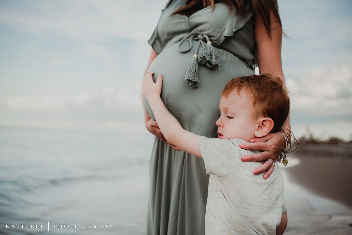 Young child hugging pregnant woman's belly on beach at sunset.