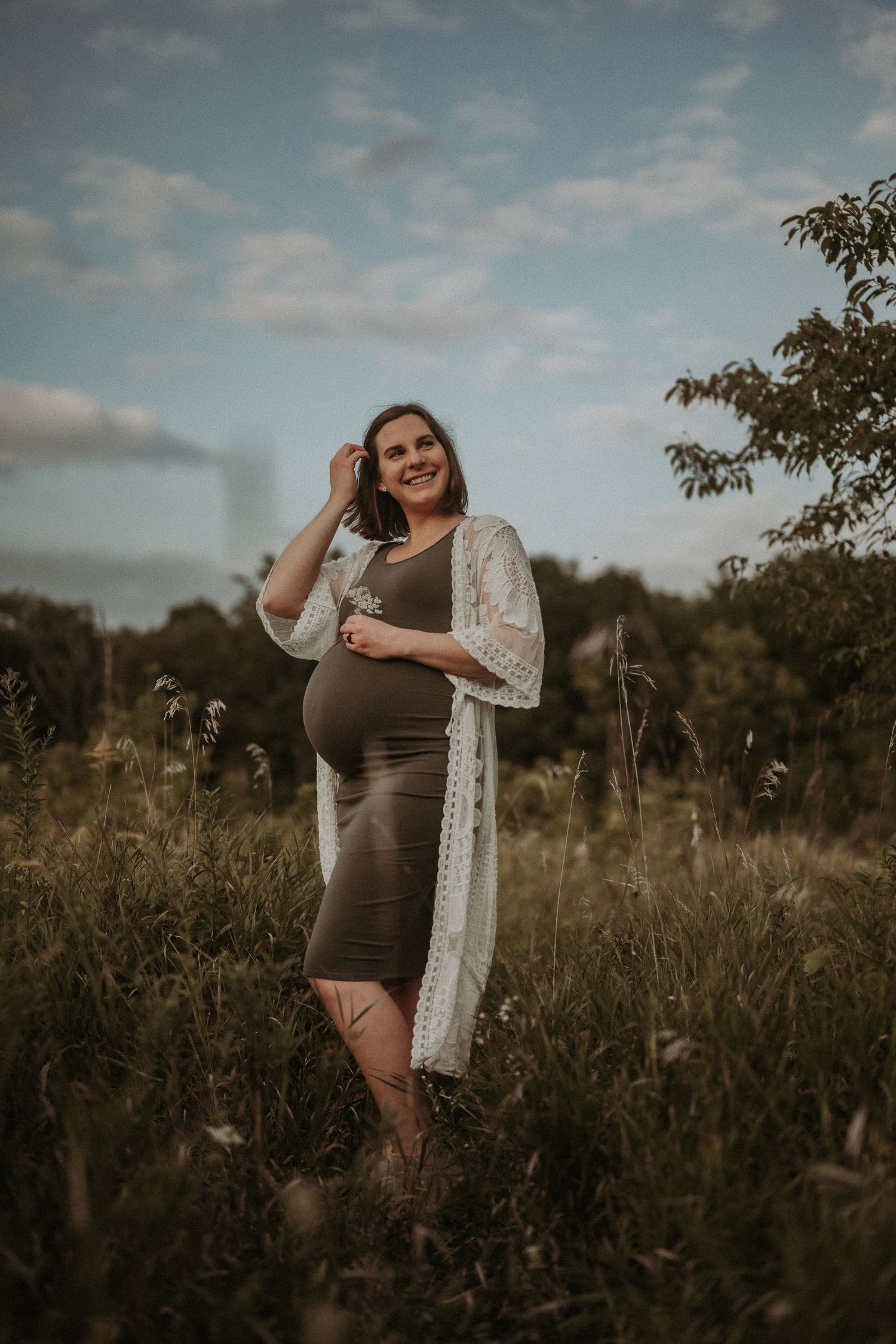 A maternity portrait in a field showing a woman in brown dress touching her hair.
