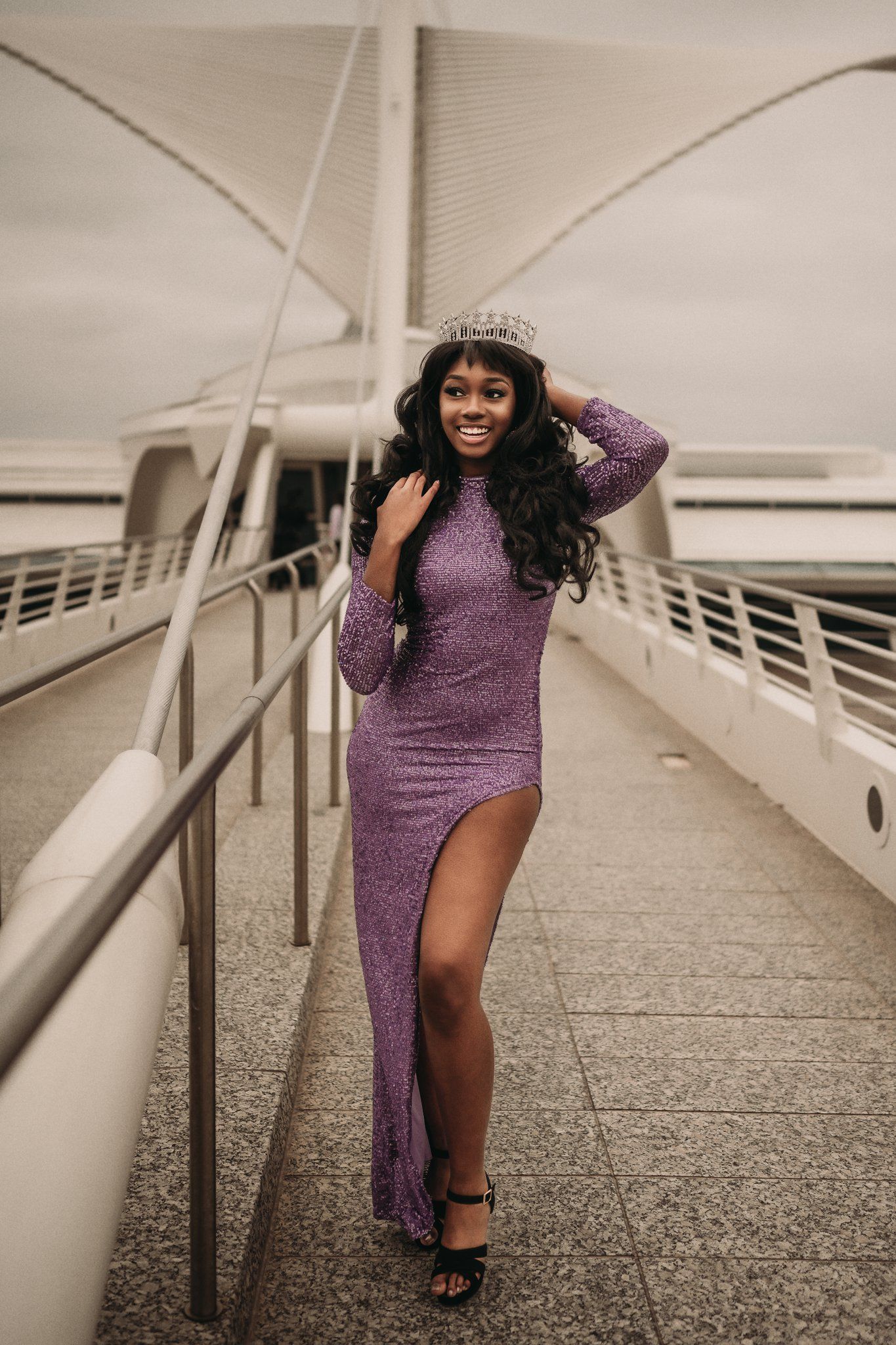 Beauty queen in glittering purple dress poses on modern bridge walkway.