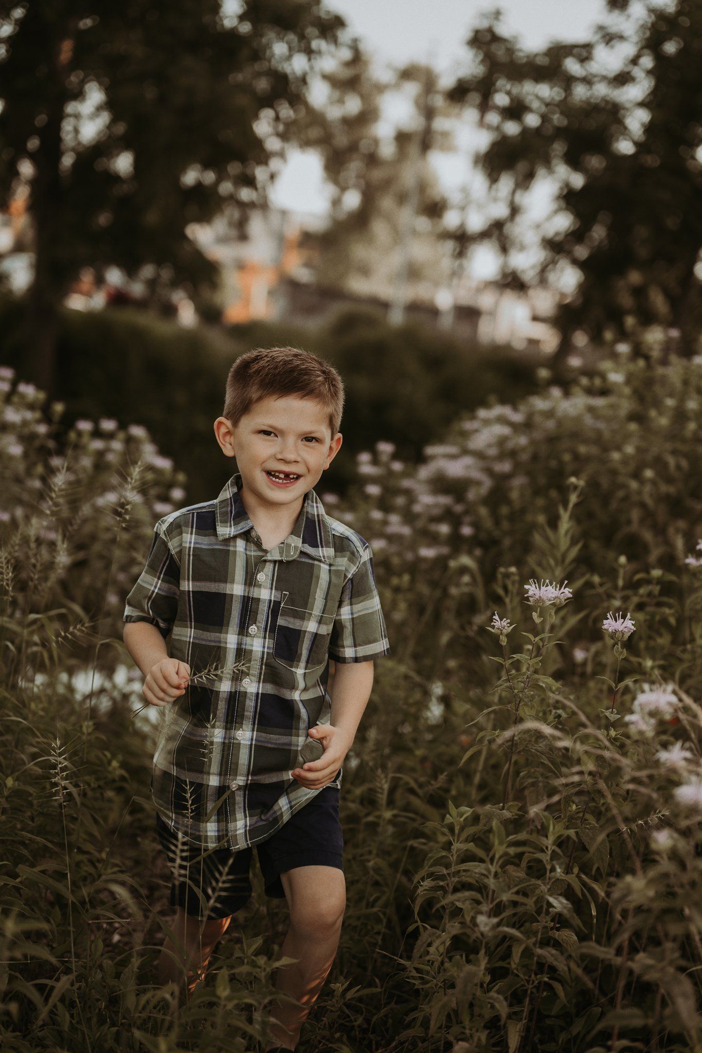 Child wearing casual plaid shirt smiles while standing among blooming wildflowers at dusk.