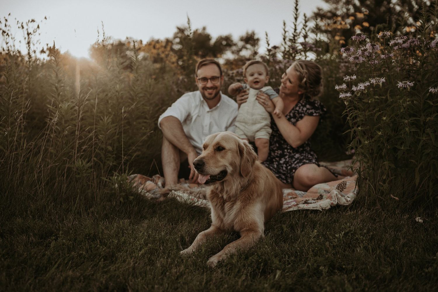 Family relaxes on blanket in wildflower field at sunset with their golden retriever.