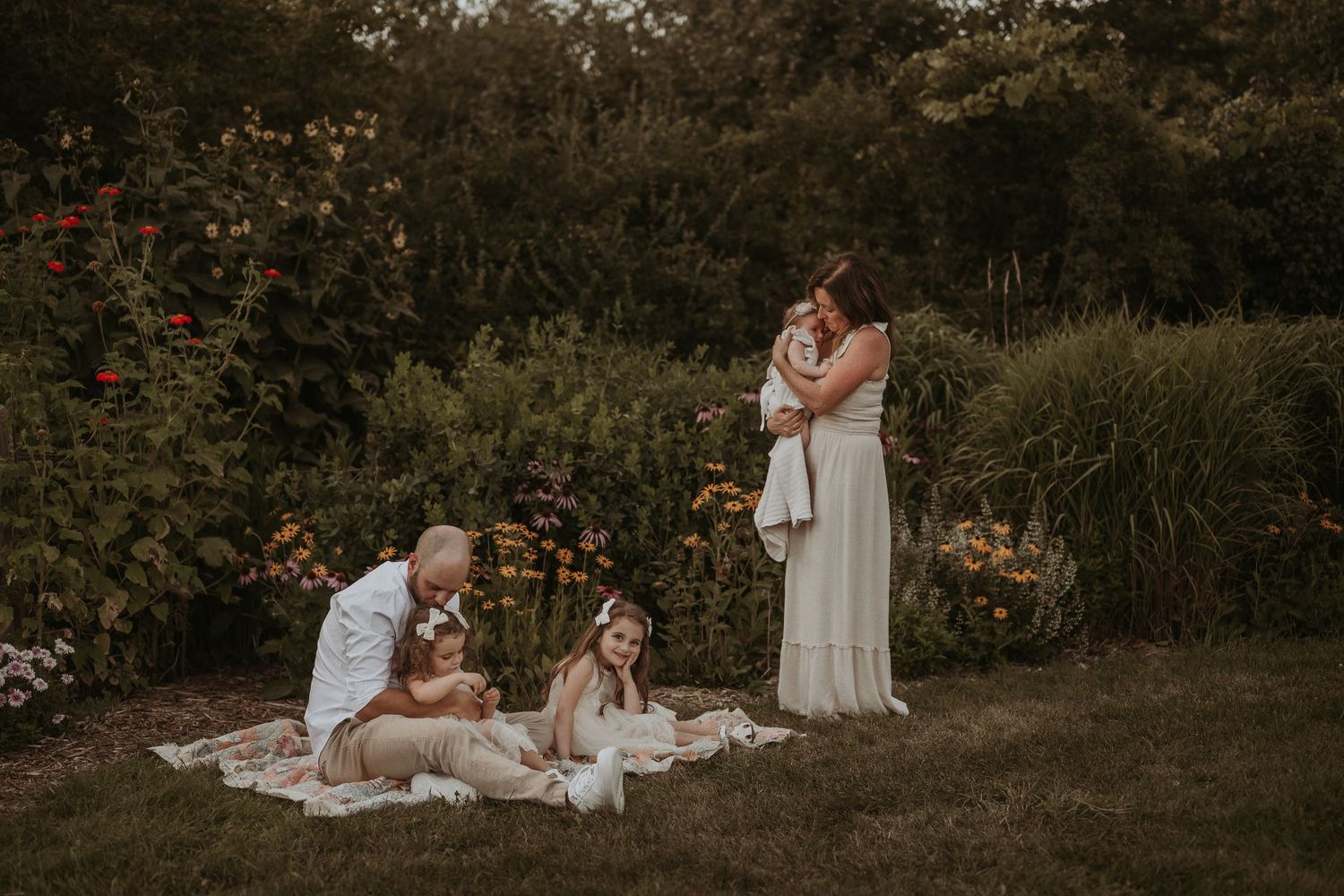 Family dressed in white relaxes on a blanket in a garden with blooming flowers at sunset.