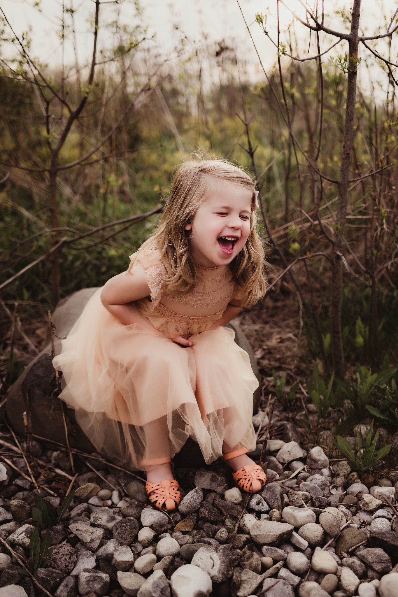 A joyful child in a peach tulle dress laughs while sitting on rocky ground.