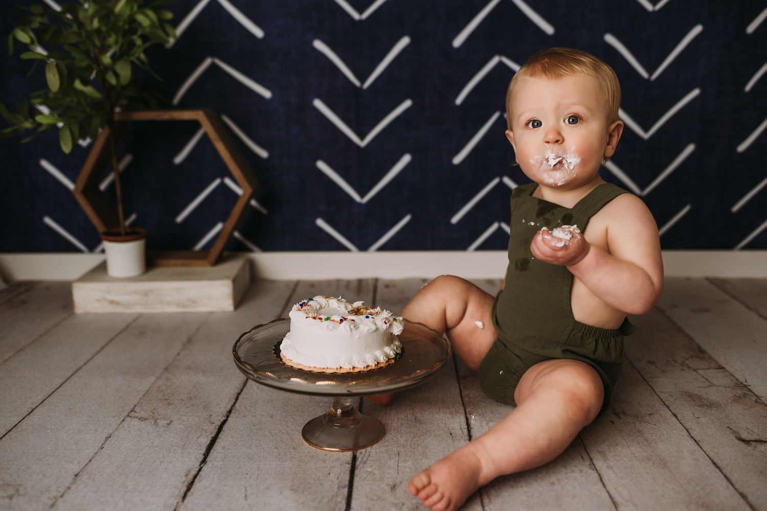 A smash cake photo setup with a white frosted cake on a pedestal against a navy patterned wall background.