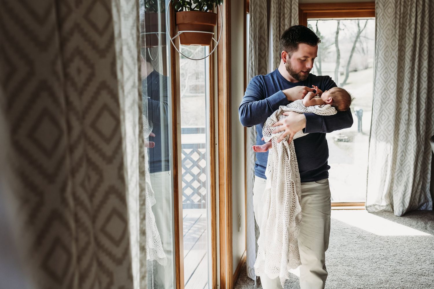 A parent holds their newborn near large windows with patterned curtains in a cozy indoor setting.