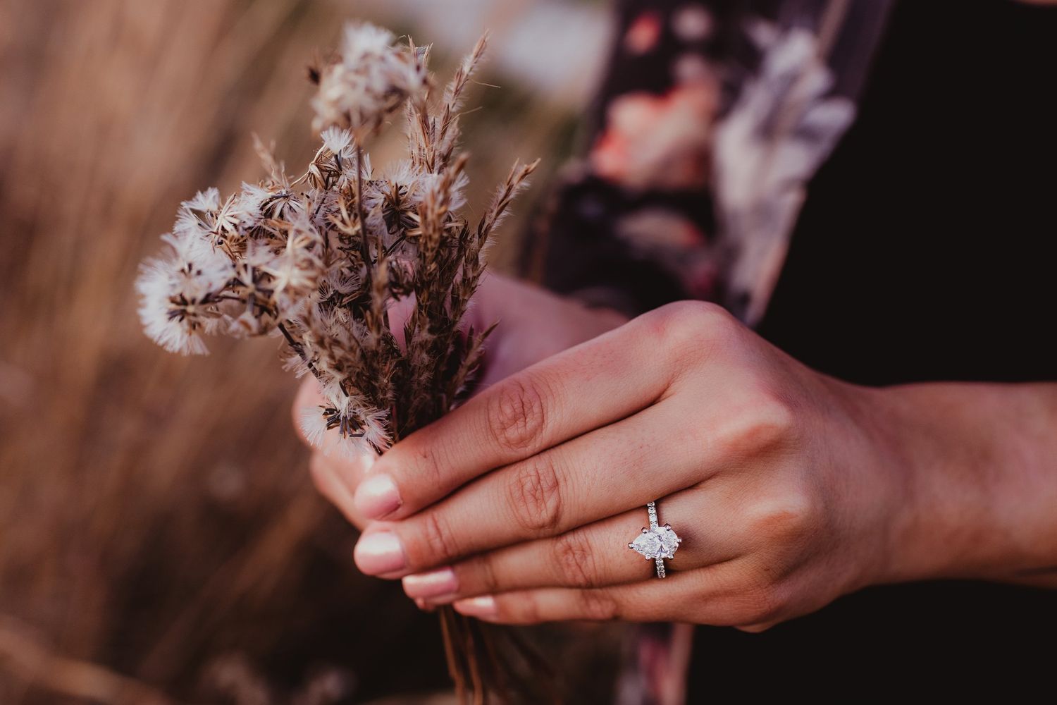 Hands hold dried wildflowers while showing off an engagement ring in warm lighting.