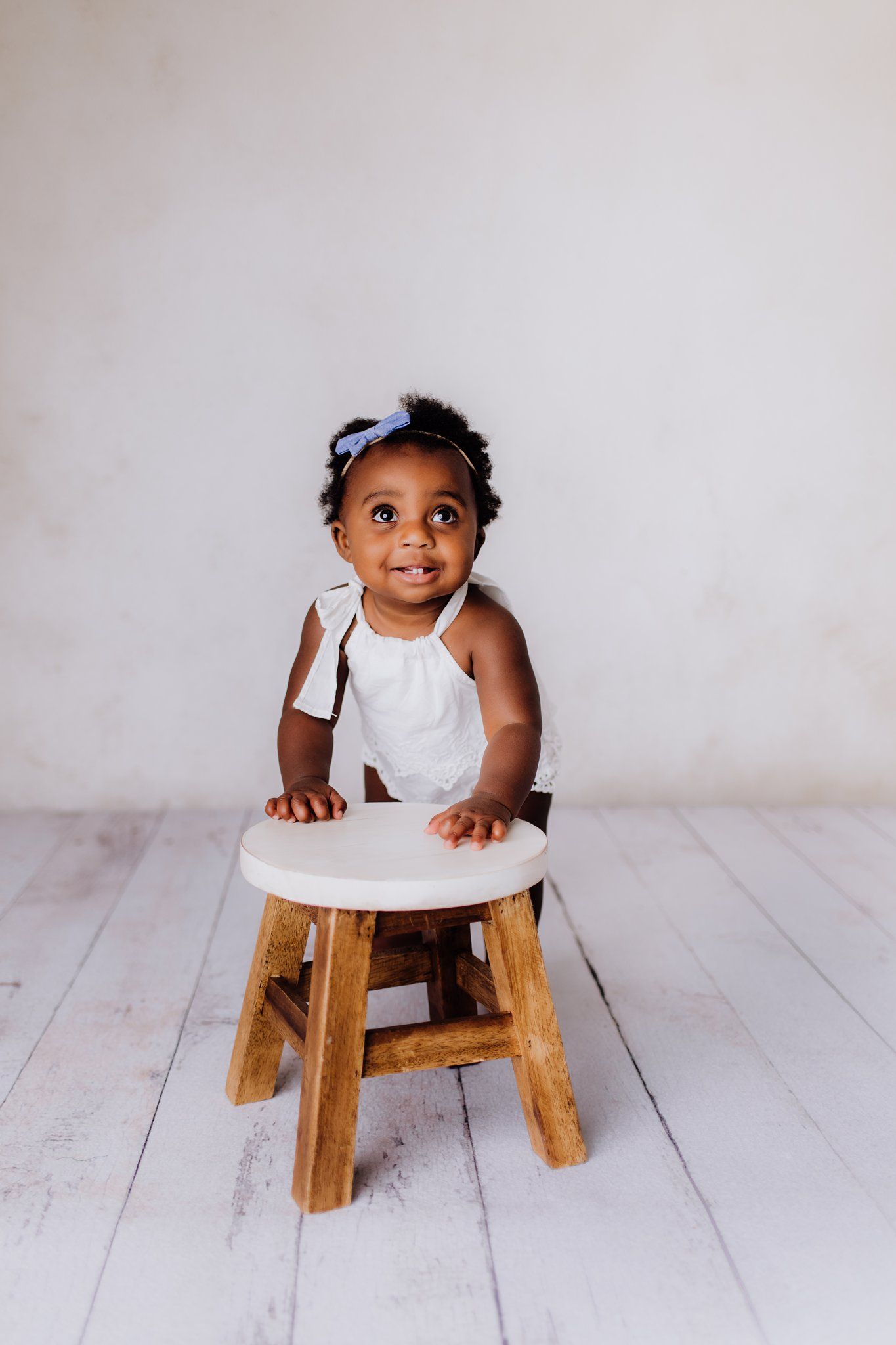 A small child in a white outfit sits on a wooden stool against a minimalist background.