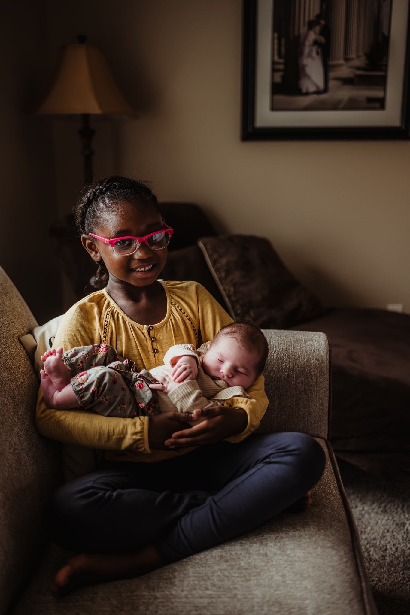Child wearing yellow shirt and glasses holds sleeping infant while sitting on brown couch.