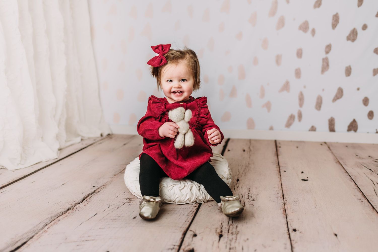 Baby in red dress smiling while holding small plush toy on wooden floor.