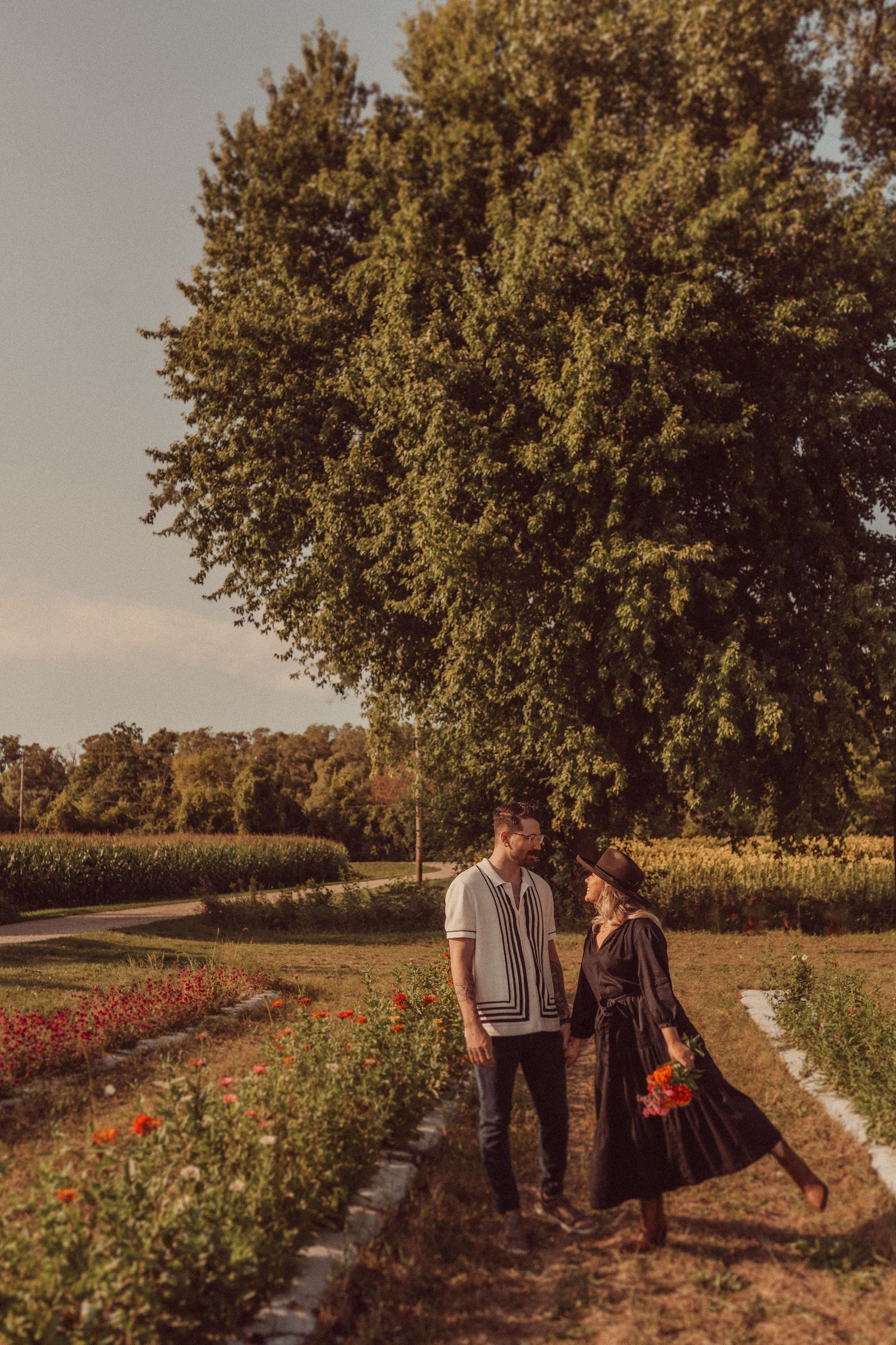 Couple strolling together on a garden path lined with flowers and tall trees.