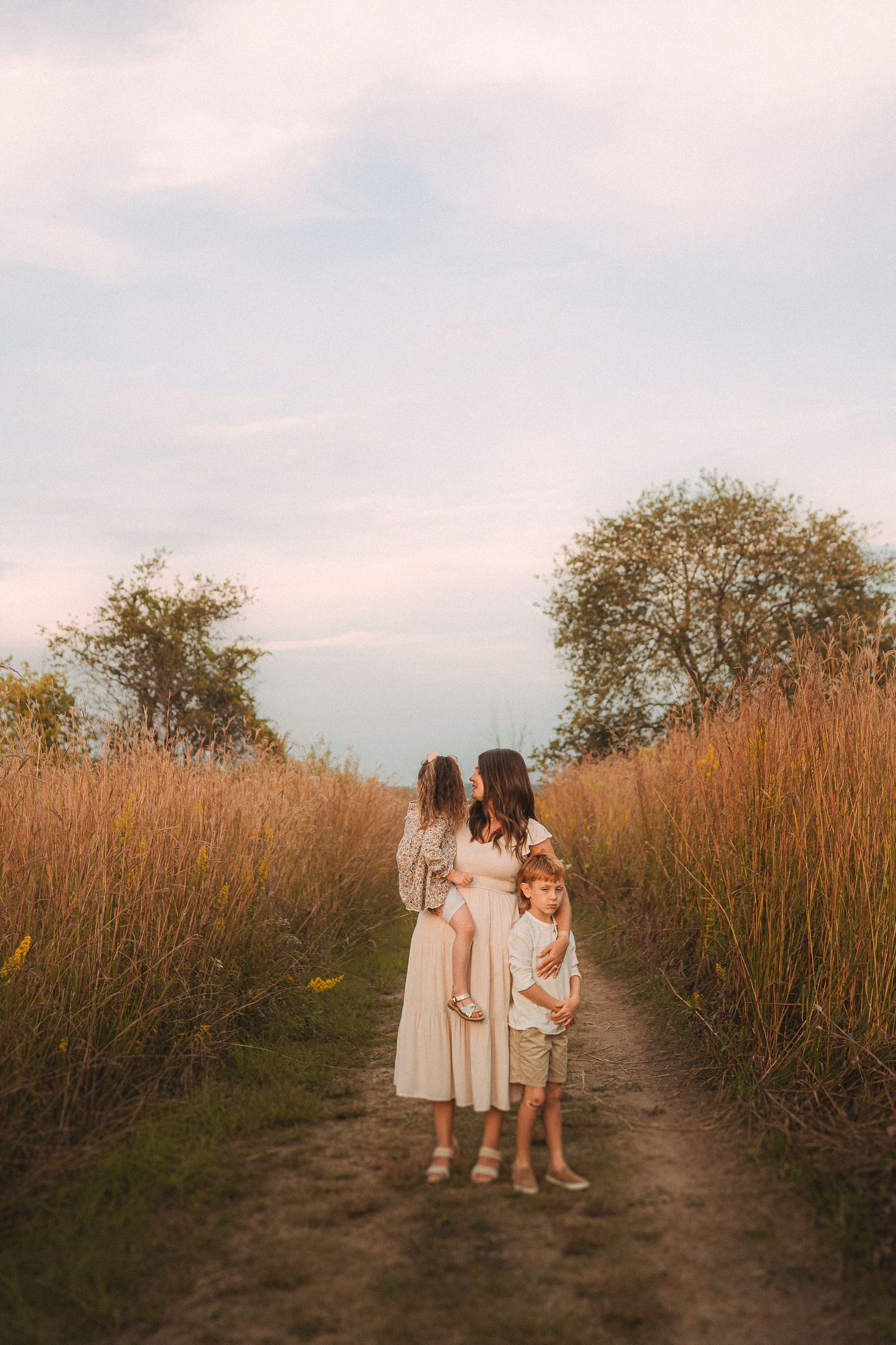 Family walking together on dirt path through tall grass at sunset.