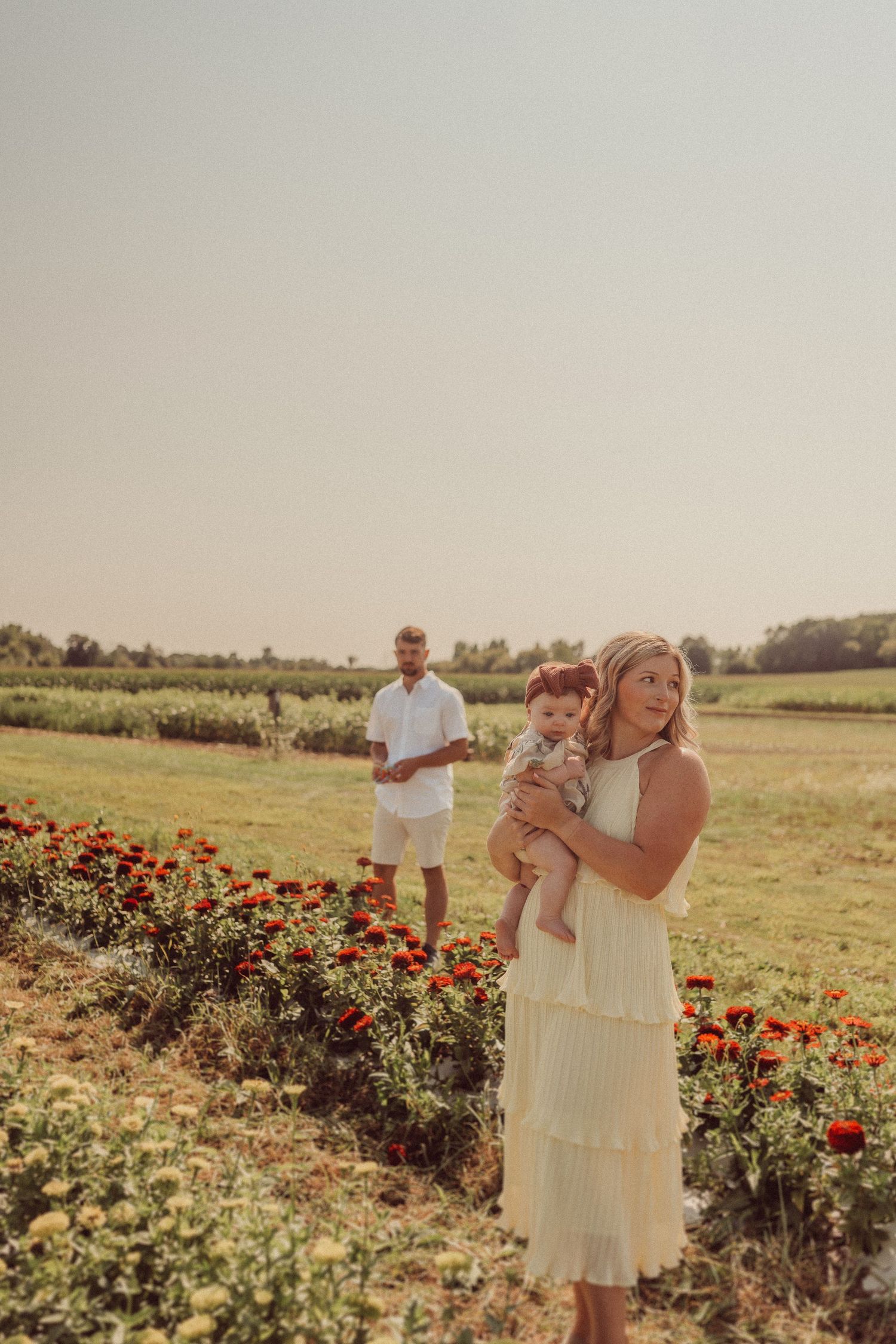 Family portrait in flower garden with mother holding baby while father stands in background.