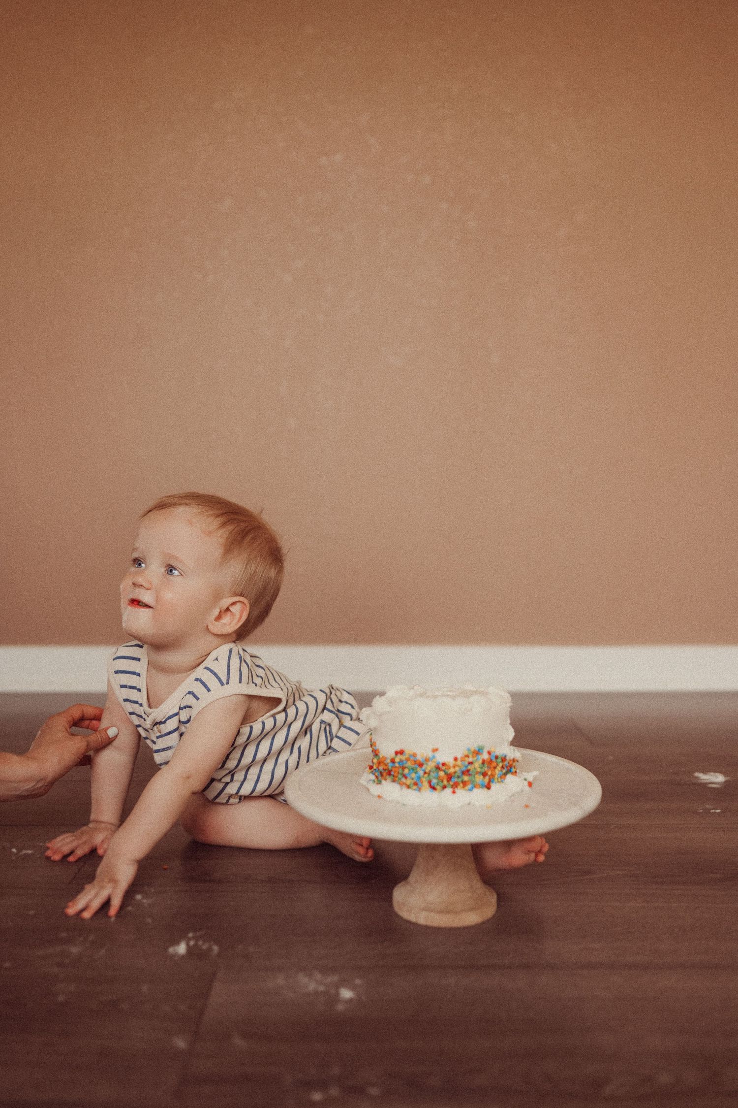 Small toddler in striped romper sitting on wooden floor next to white cake on pedestal with colorful sprinkles.