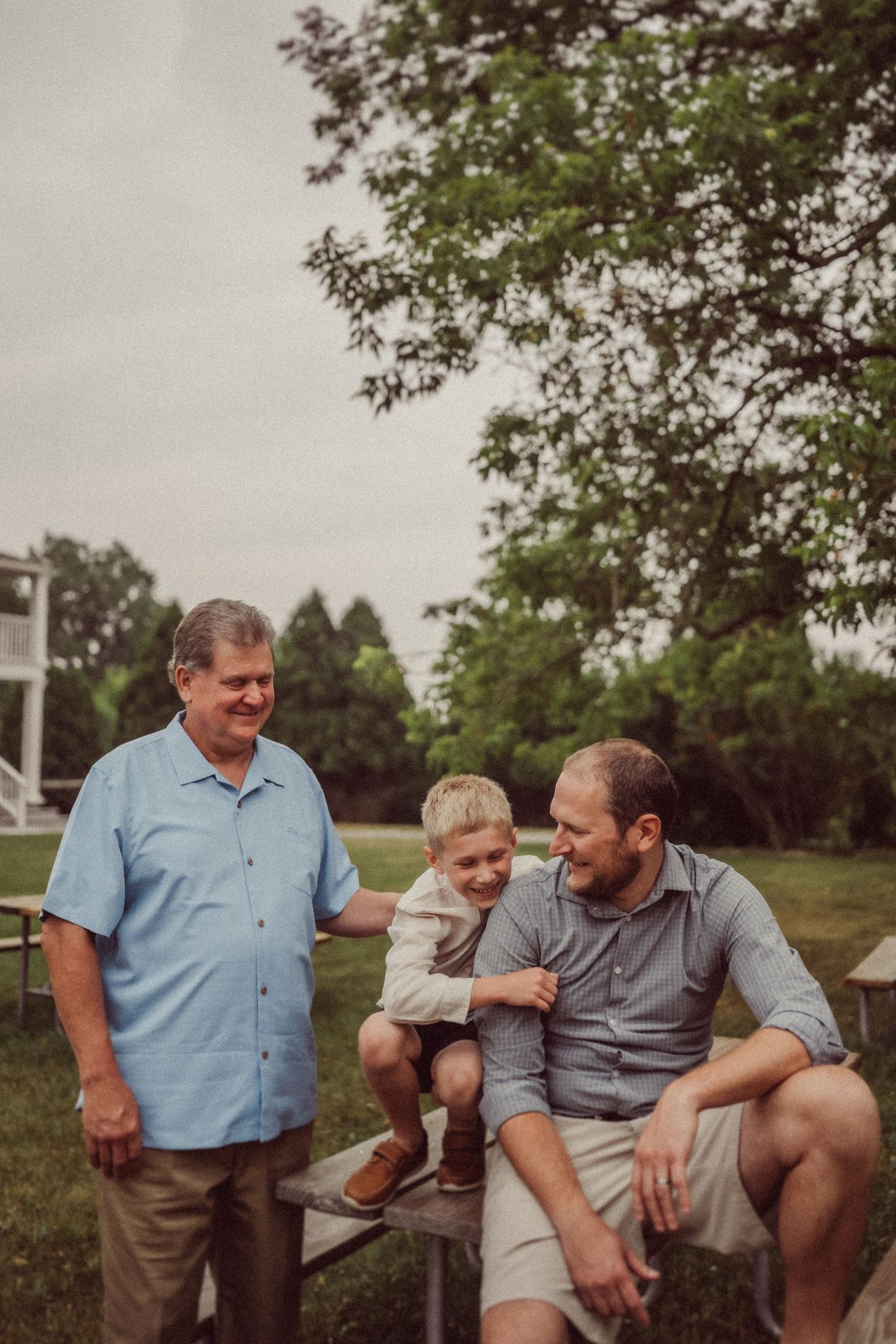 Three generations gather on wooden bleachers outdoors on a cloudy day.