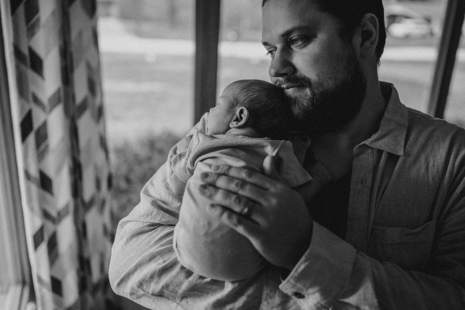 Black and white image of adult holding sleeping infant by window with curtains.