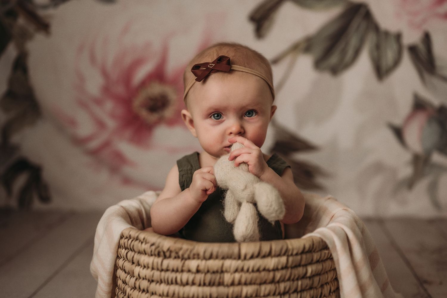Baby wearing dark romper sits in woven basket against floral backdrop.