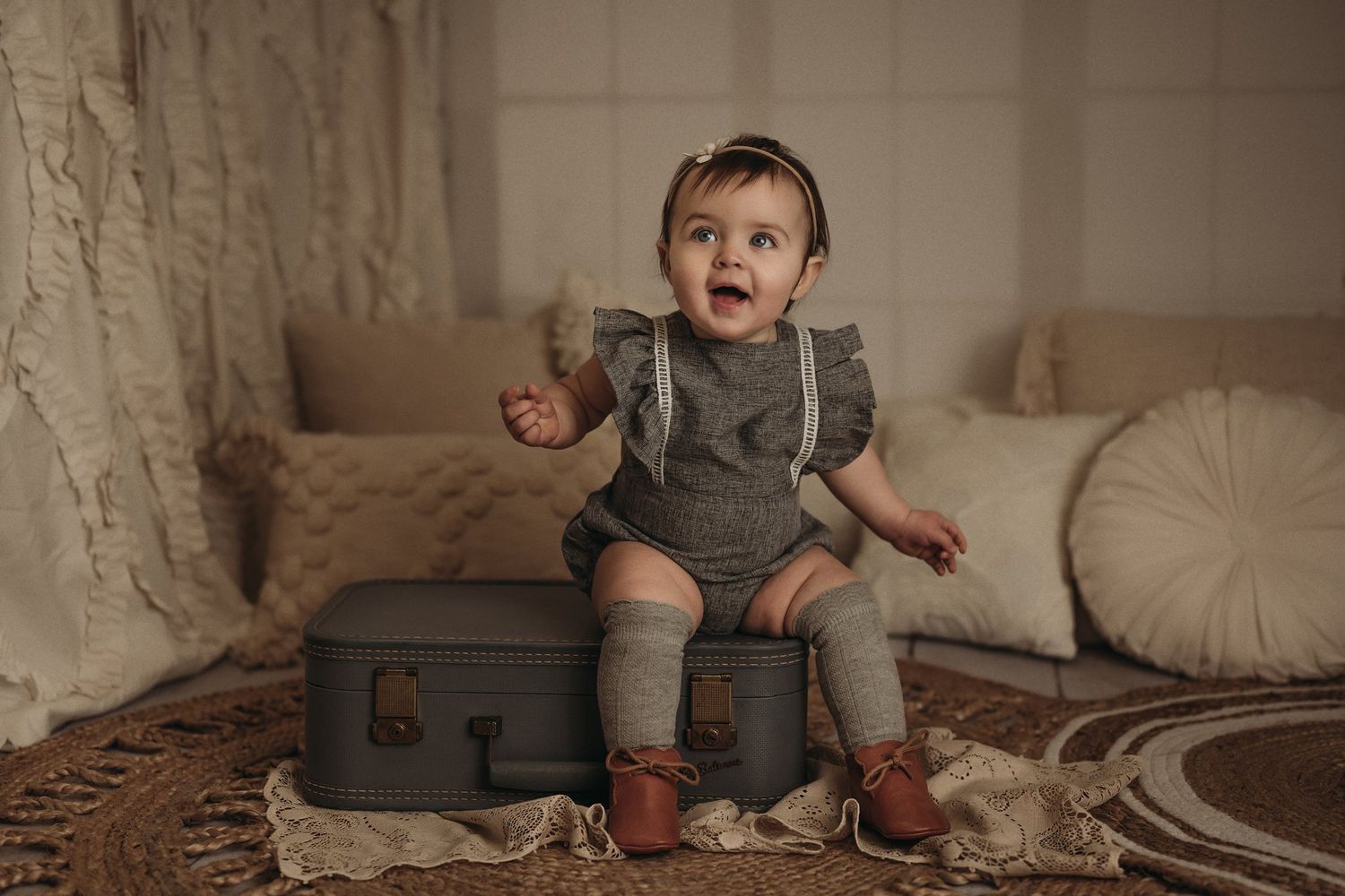 Baby in grey outfit with ruffles sits on vintage luggage against neutral backdrop.