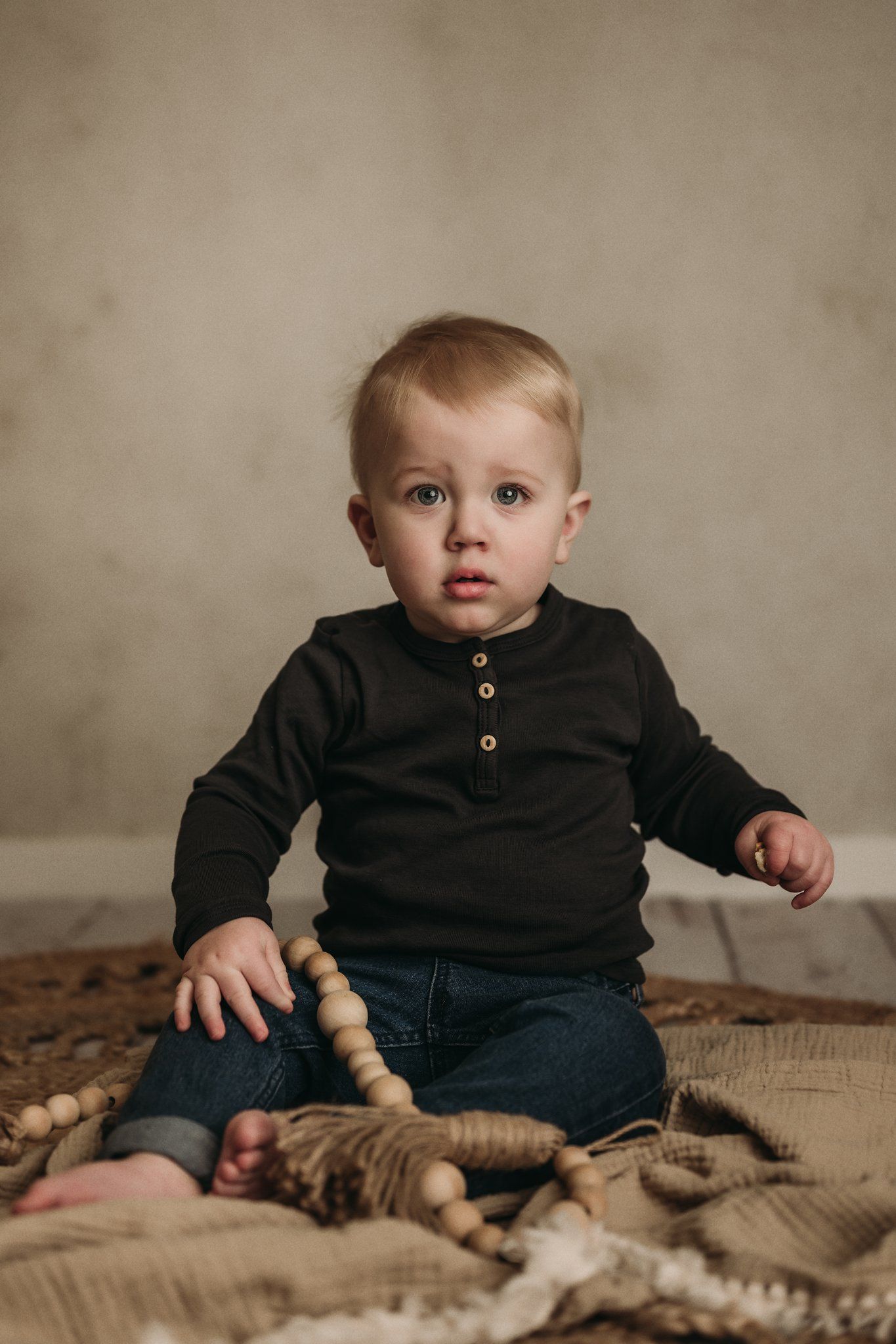 Studio portrait of a toddler wearing a black henley shirt and jeans sitting on textured fabric.