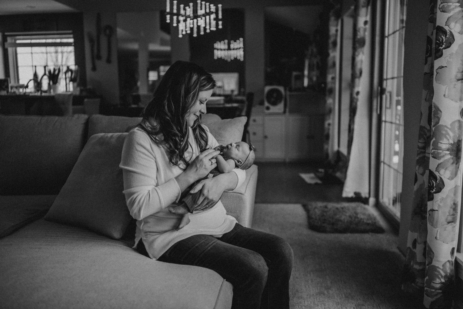Black and white portrait of tender moment between parent and infant on sofa near window.