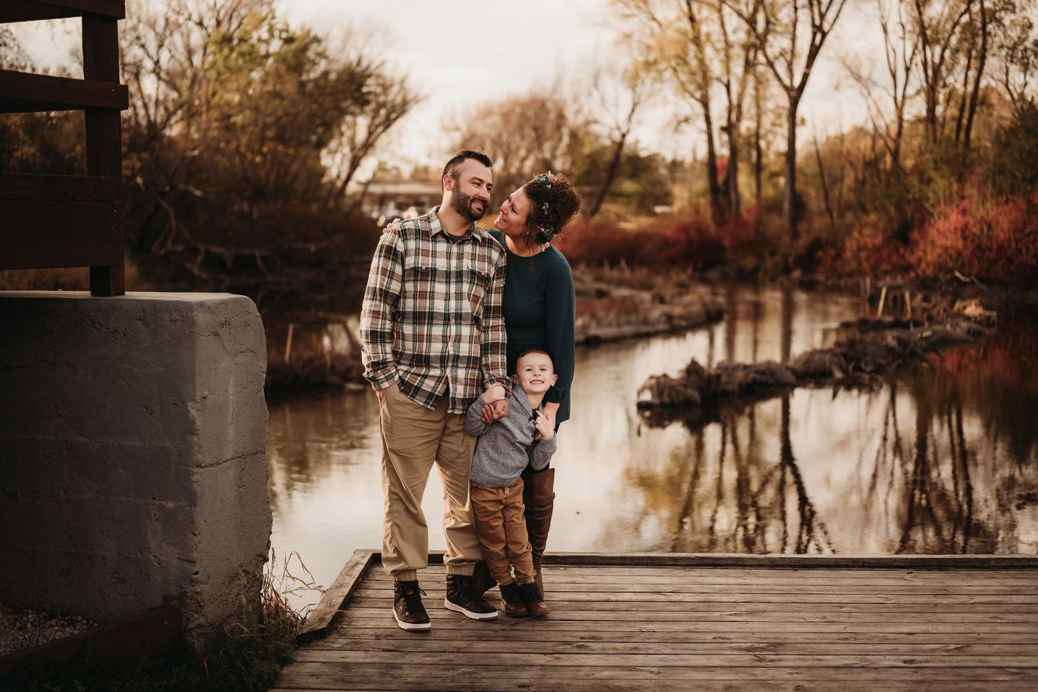Family standing together on wooden dock by tranquil pond in autumn setting.