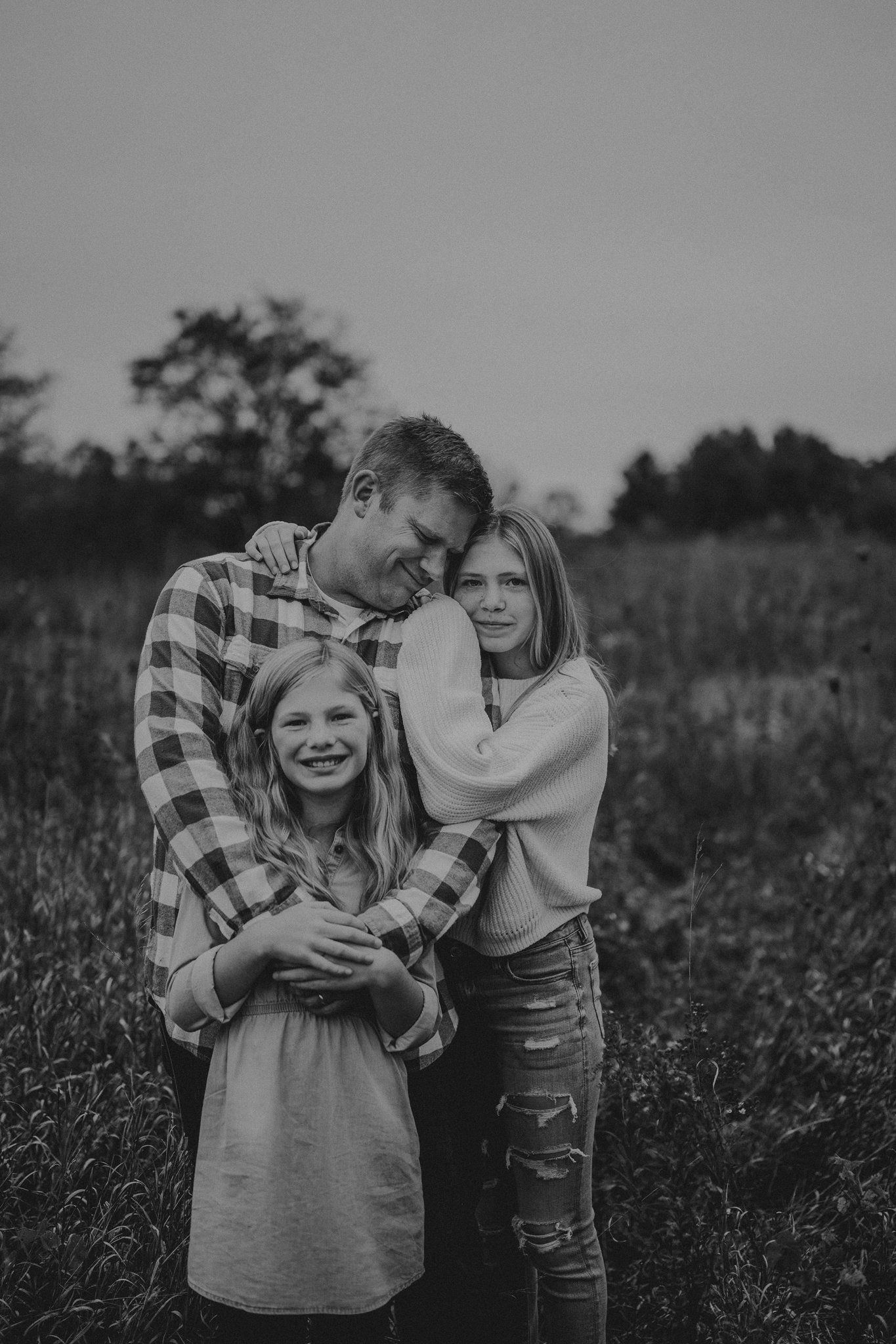 Black and white portrait of family embracing in a rustic field at dusk.