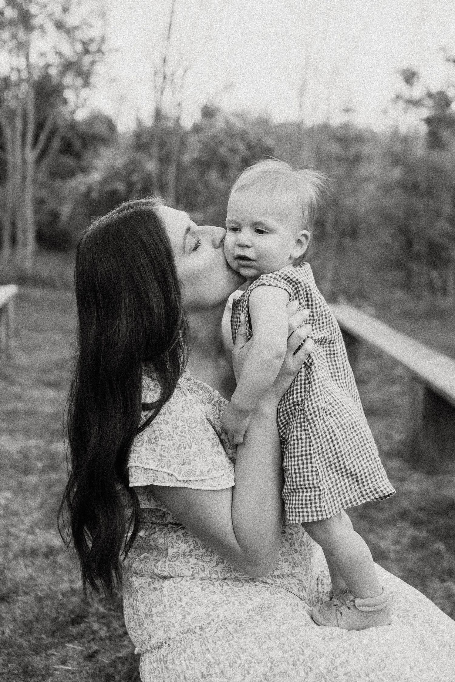 Mother kissing her baby girl on the cheek while holding her outdoors in a black and white portrait.