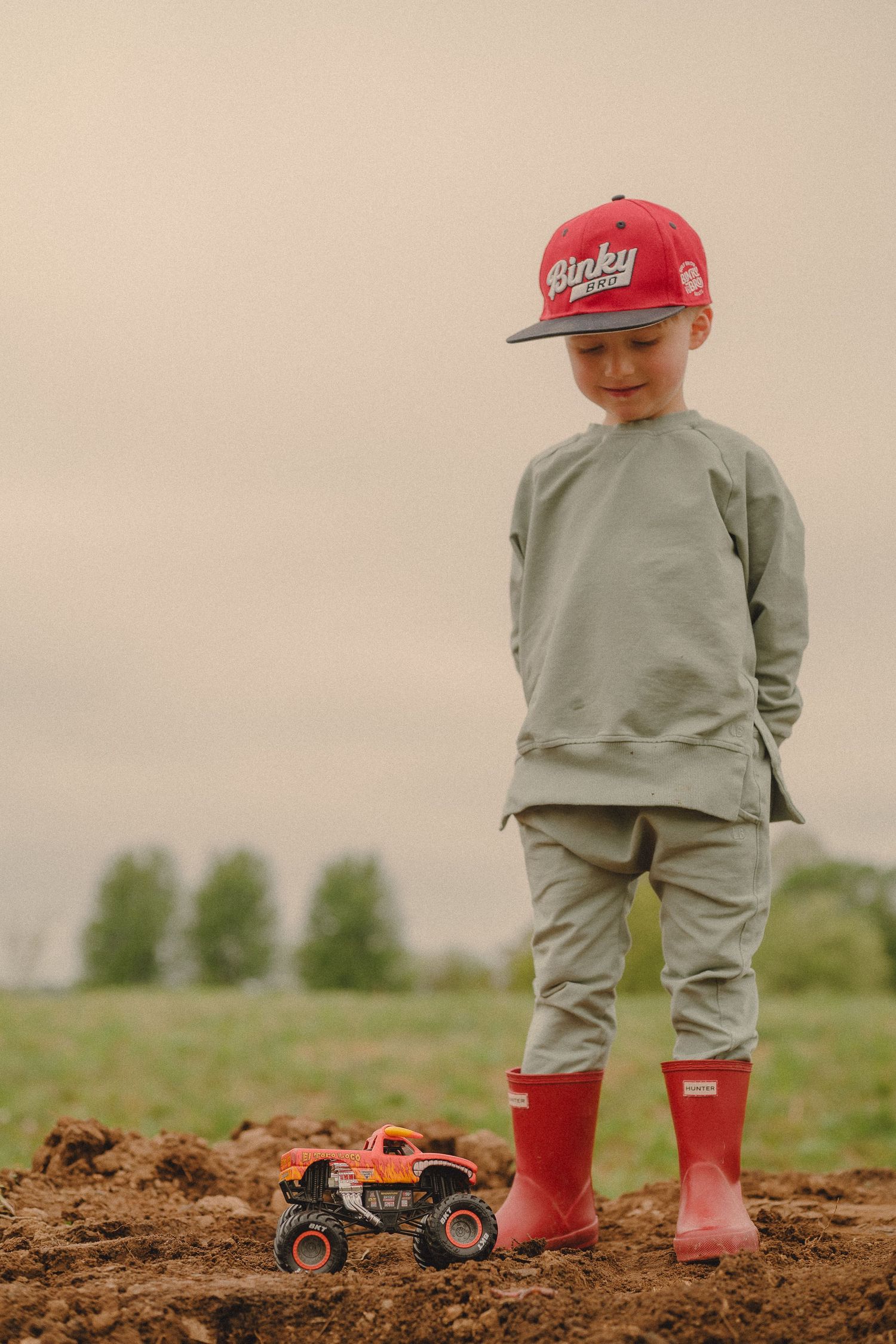 Young boy in red cap and boots looks down at toy monster truck on dirt field.