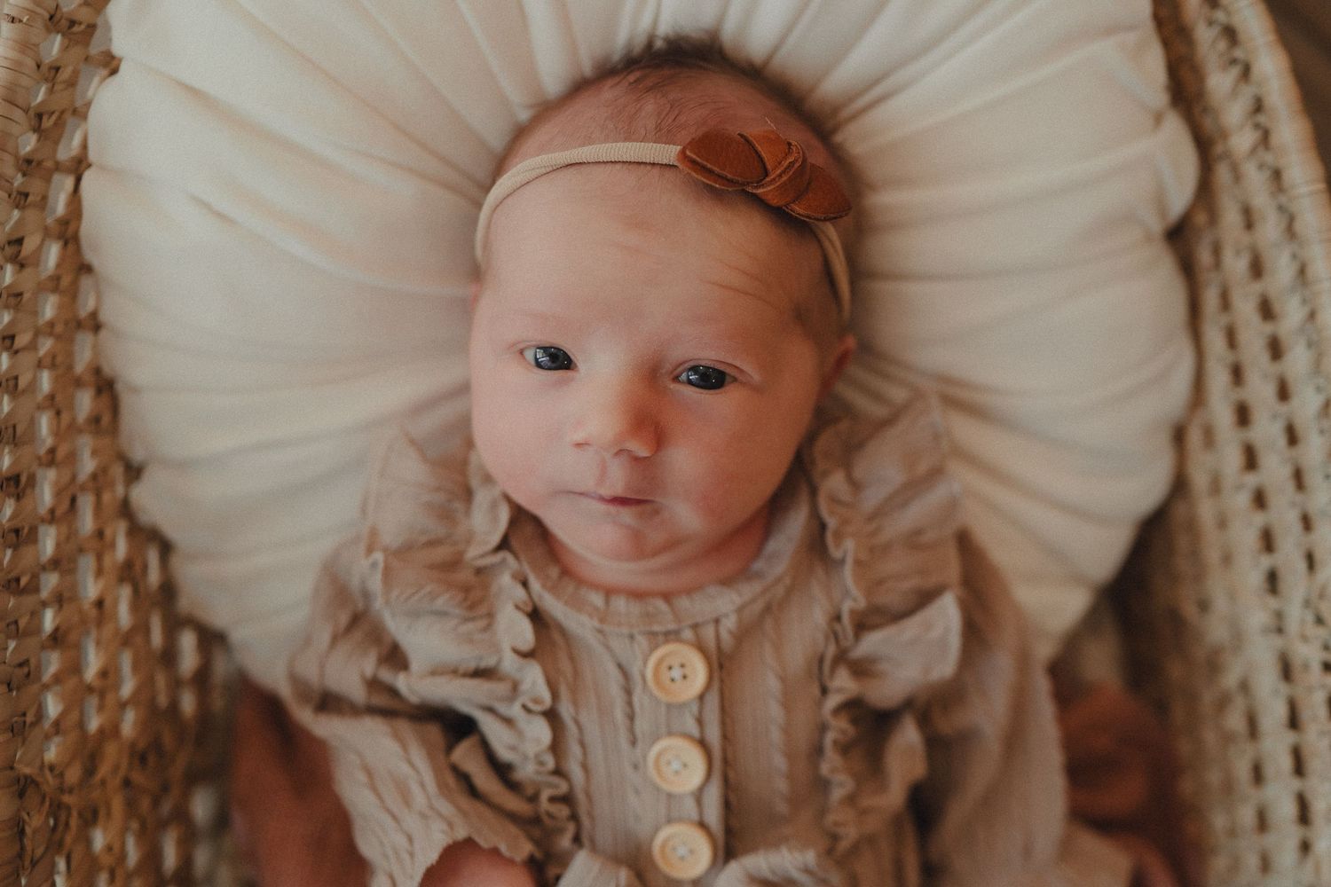 Baby wearing a knitted brown hat and white onesie lies on a white blanket.