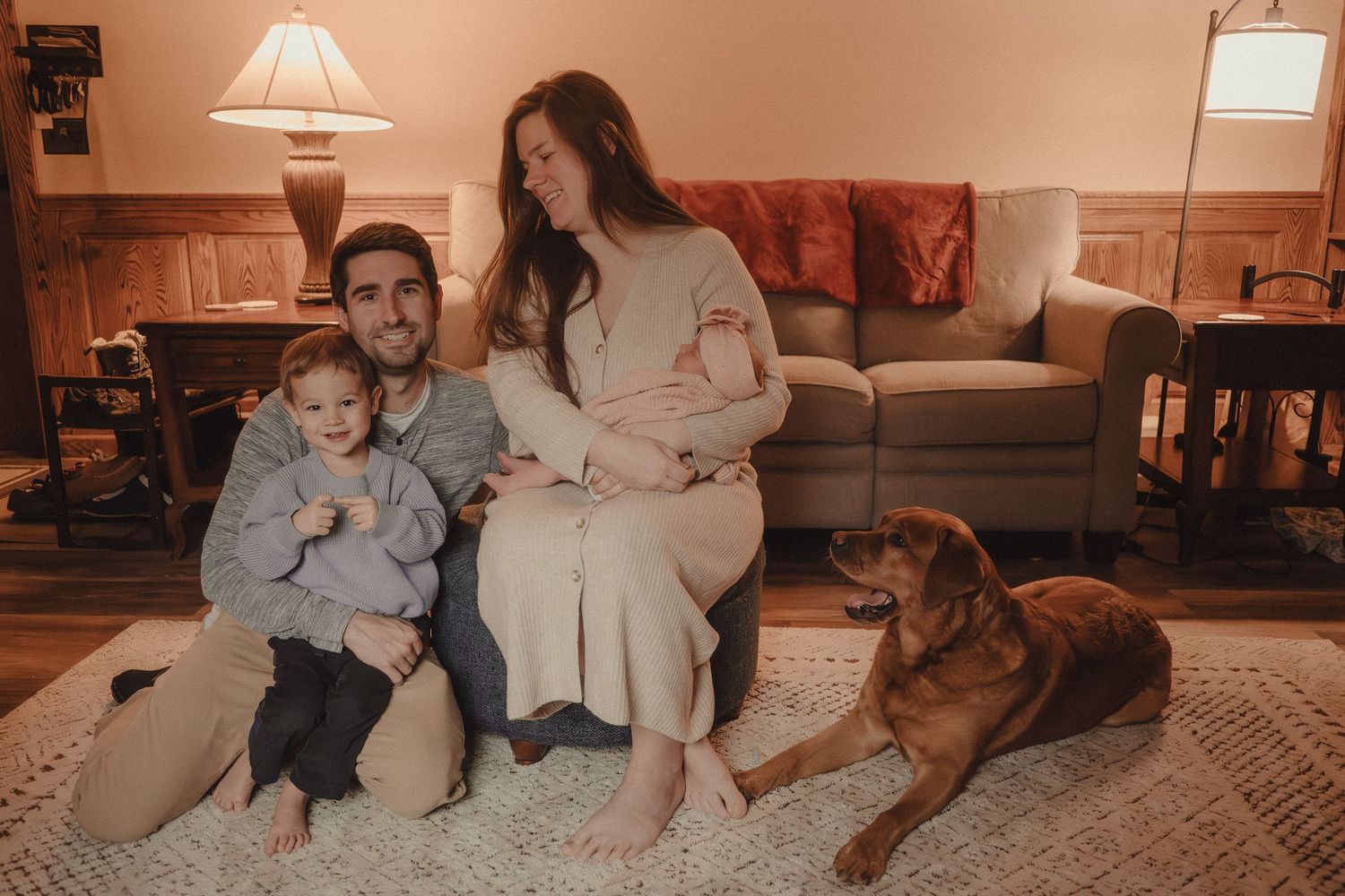 A family relaxes together in their cozy living room with a brown dog on a carpeted floor at dusk.