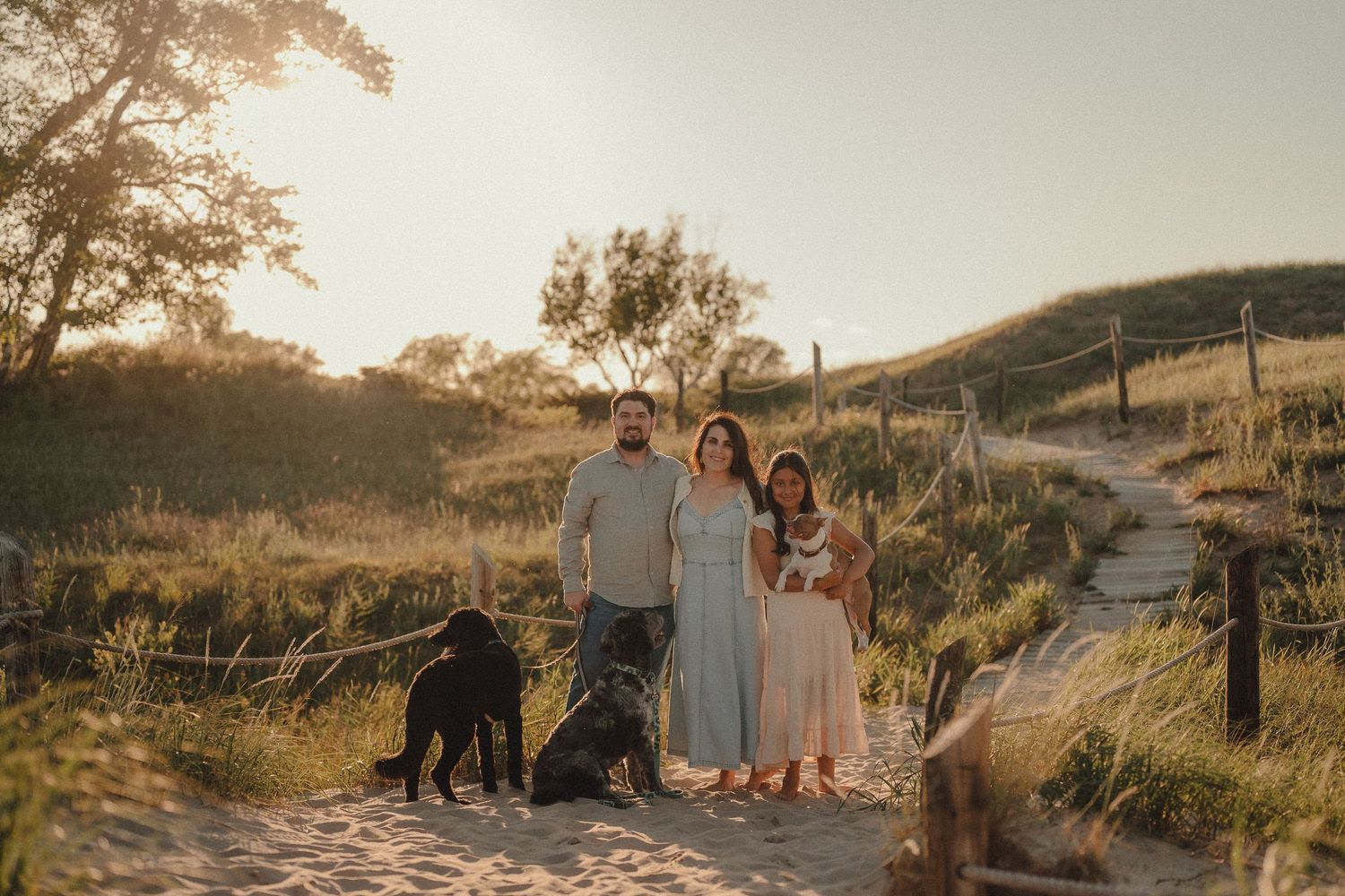 Family with two black dogs posing on sandy beach path surrounded by golden grass at sunset.