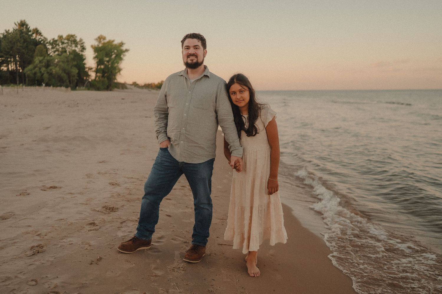 Couple standing together on a sandy beach at sunset with calm water and trees in the background.