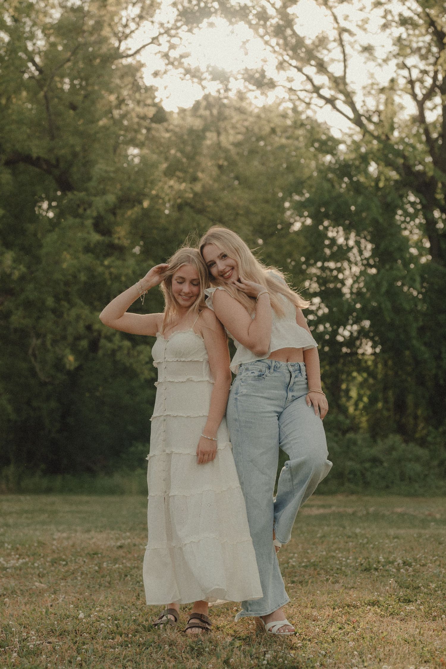 Two blonde women in bohemian outfits pose together in a lush green outdoor setting during golden hour.
