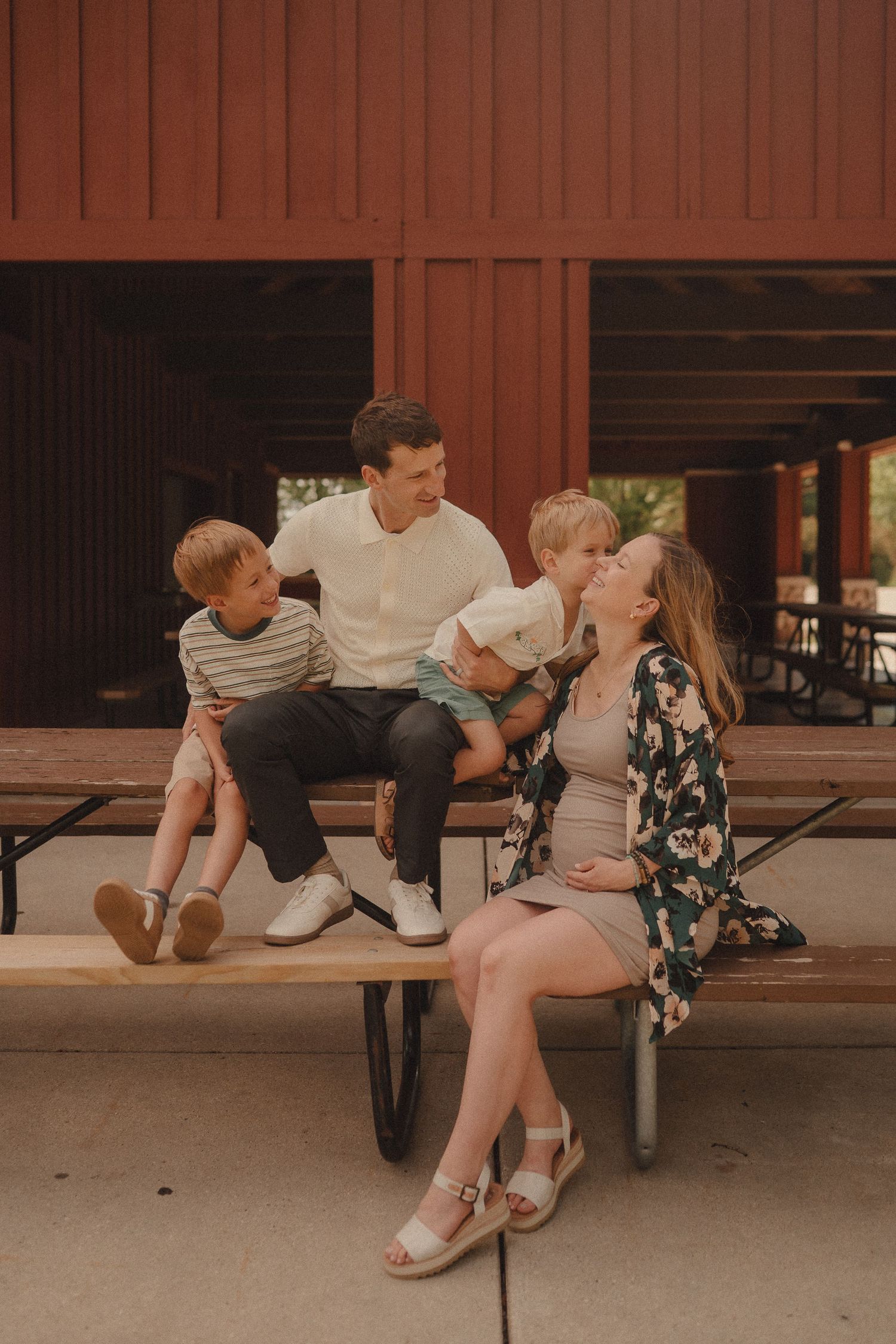 Happy family of four sitting on a picnic bench near a red barn, mom visibly pregnant.