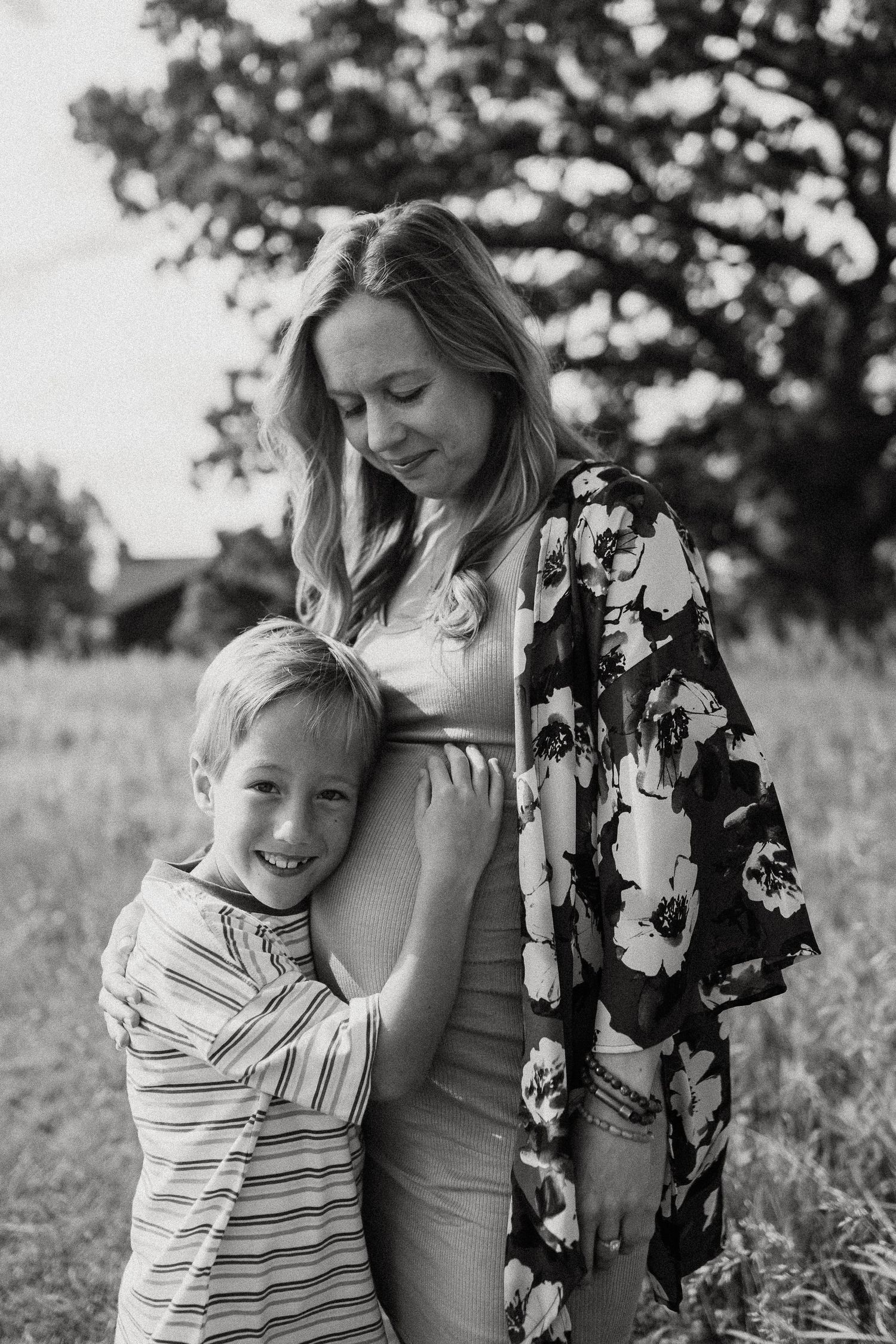 Black and white photo of a smiling boy hugging his pregnant mothers belly outdoors.