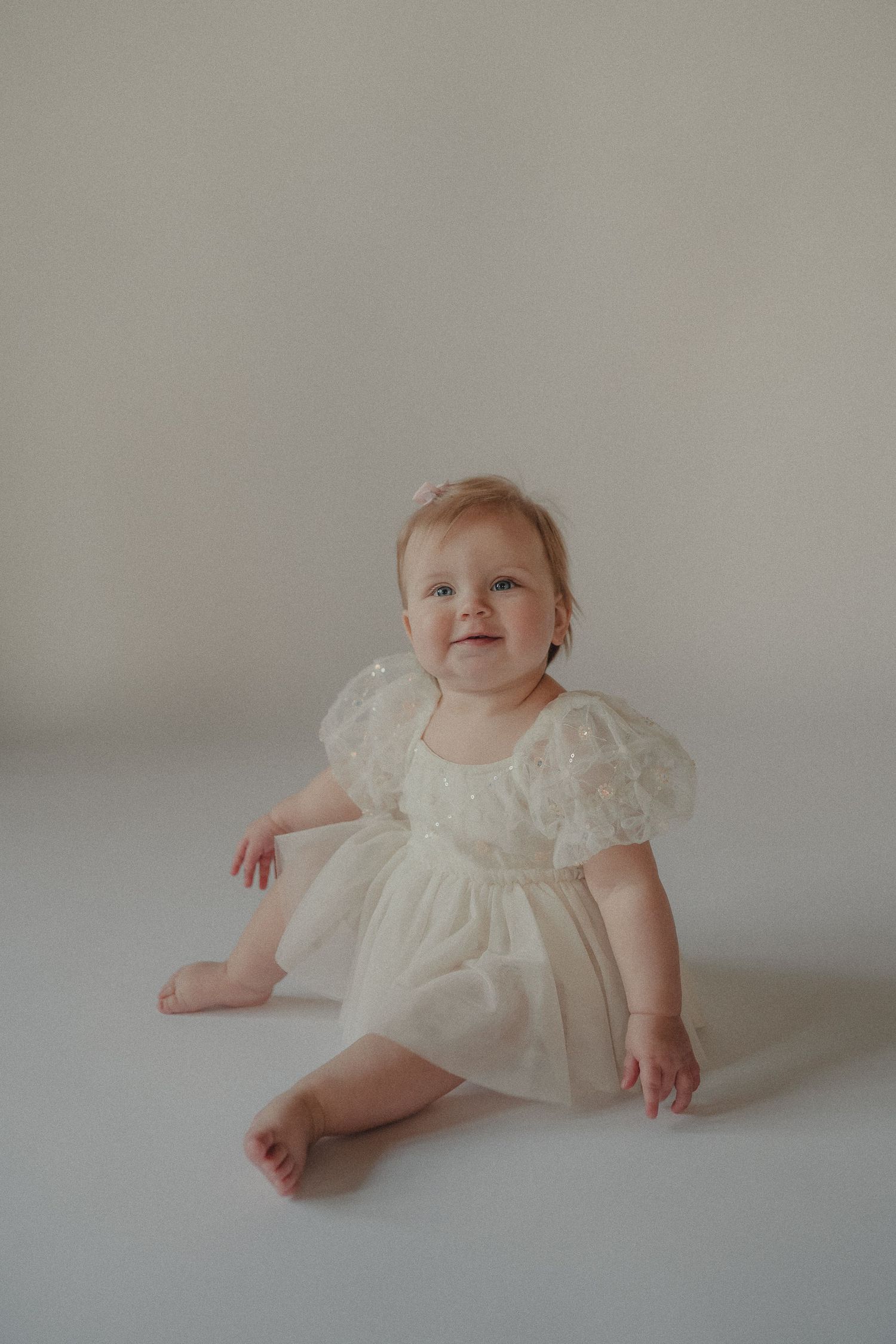 Sweet baby girl in ivory tulle dress sits barefoot smiling during first birthday milestone photo session.