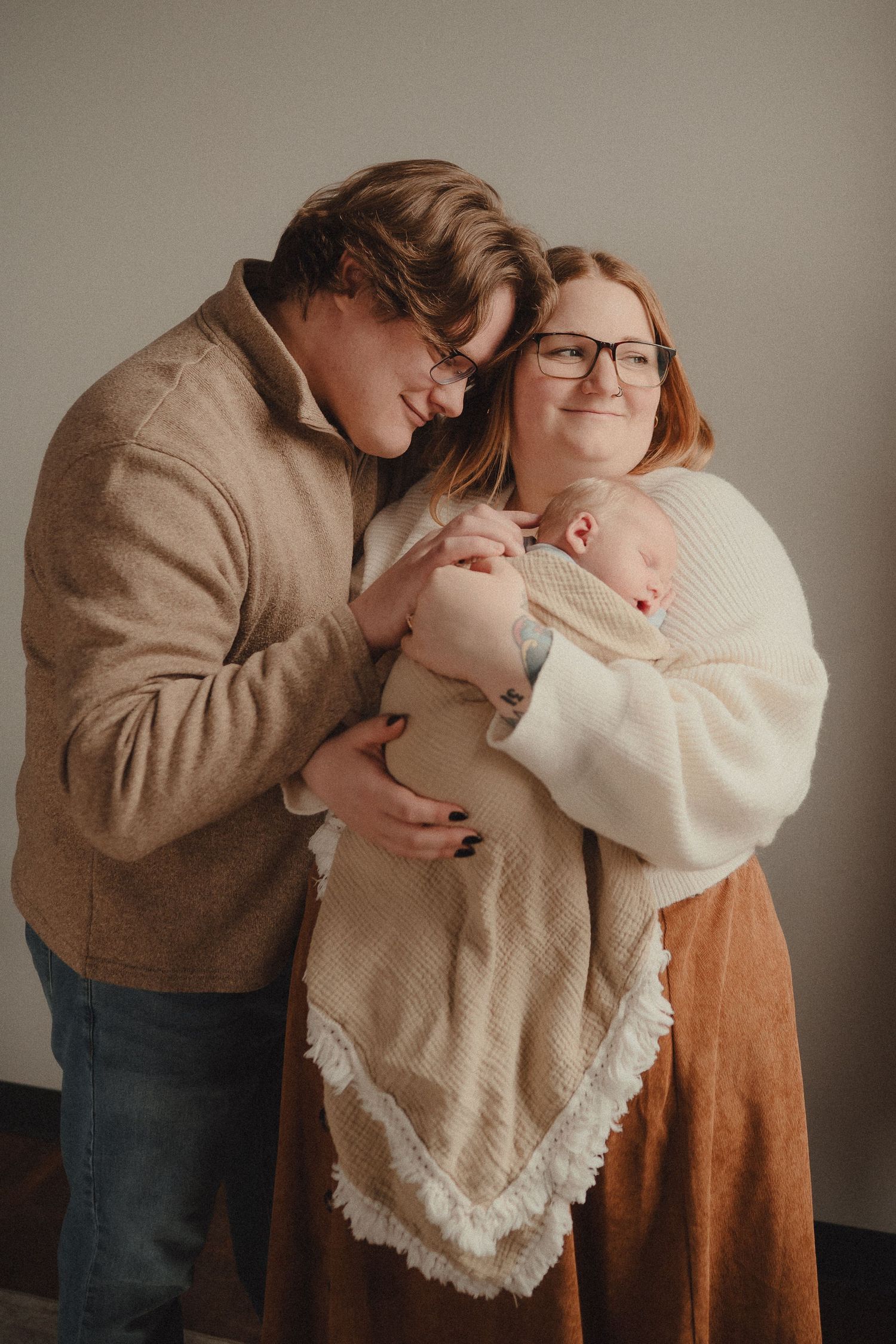 A couple in neutral tones lovingly holds their newborn baby wrapped in a cream blanket during a studio portrait session.