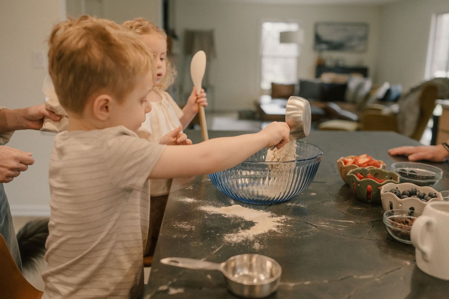 Two young children use a wooden spoon and strainer to mix ingredients on a flour-covered kitchen counter.