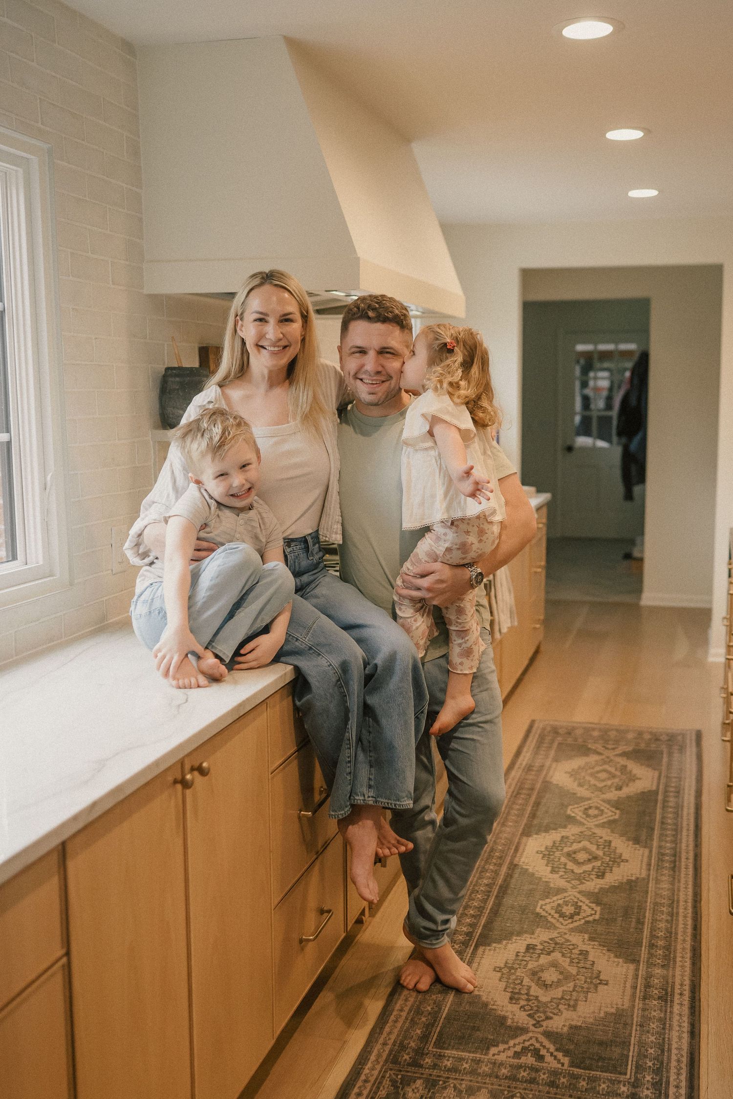 Happy family of four posing in a modern kitchen with wood cabinets, marble countertops, and hardwood floors.