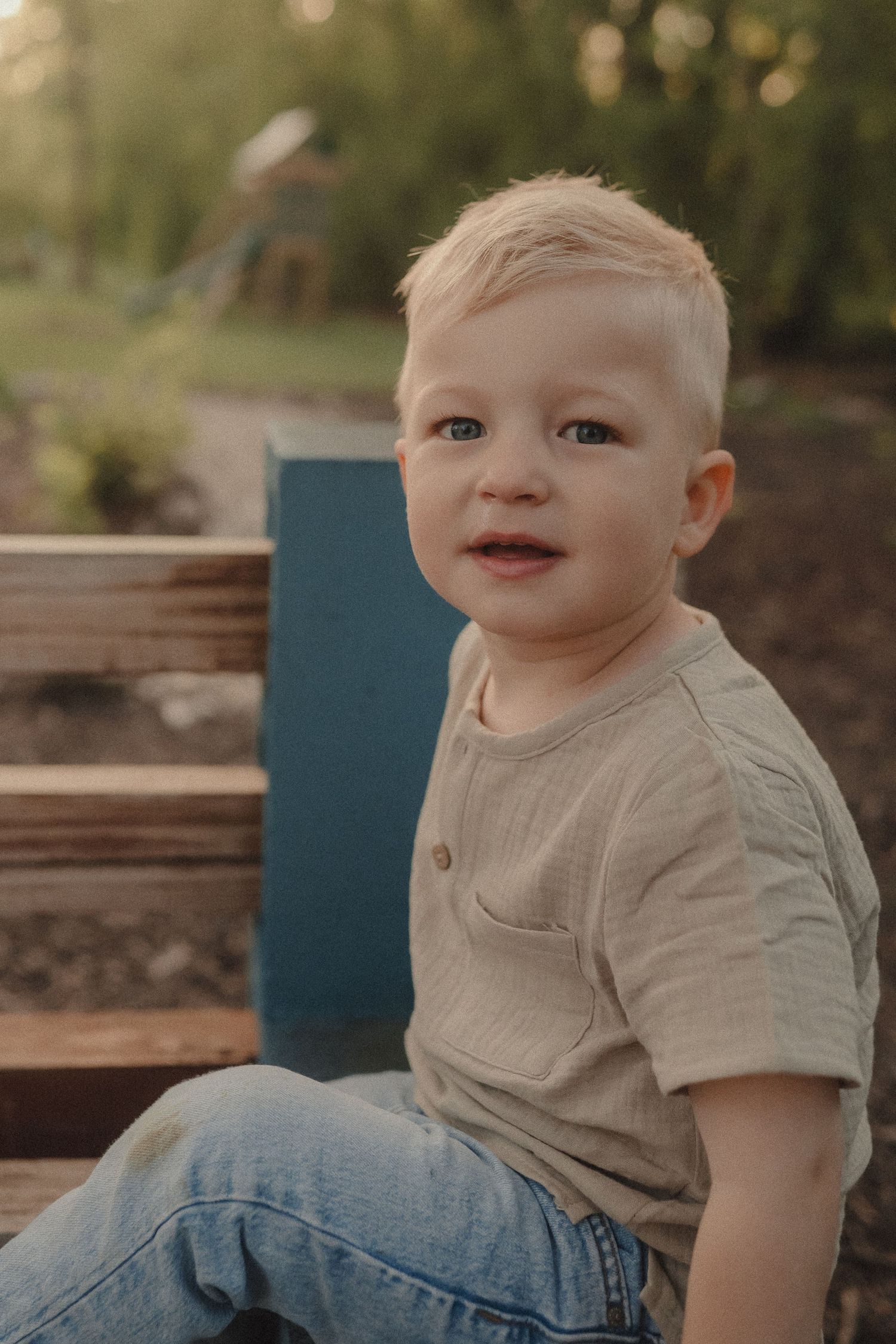 Cute blond toddler boy sitting on wooden bench outdoors in golden sunlight.