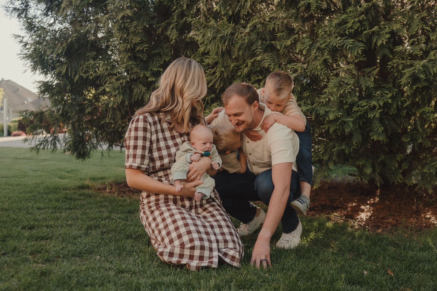 Family with two young children and newborn playing together on green grass near evergreen trees.