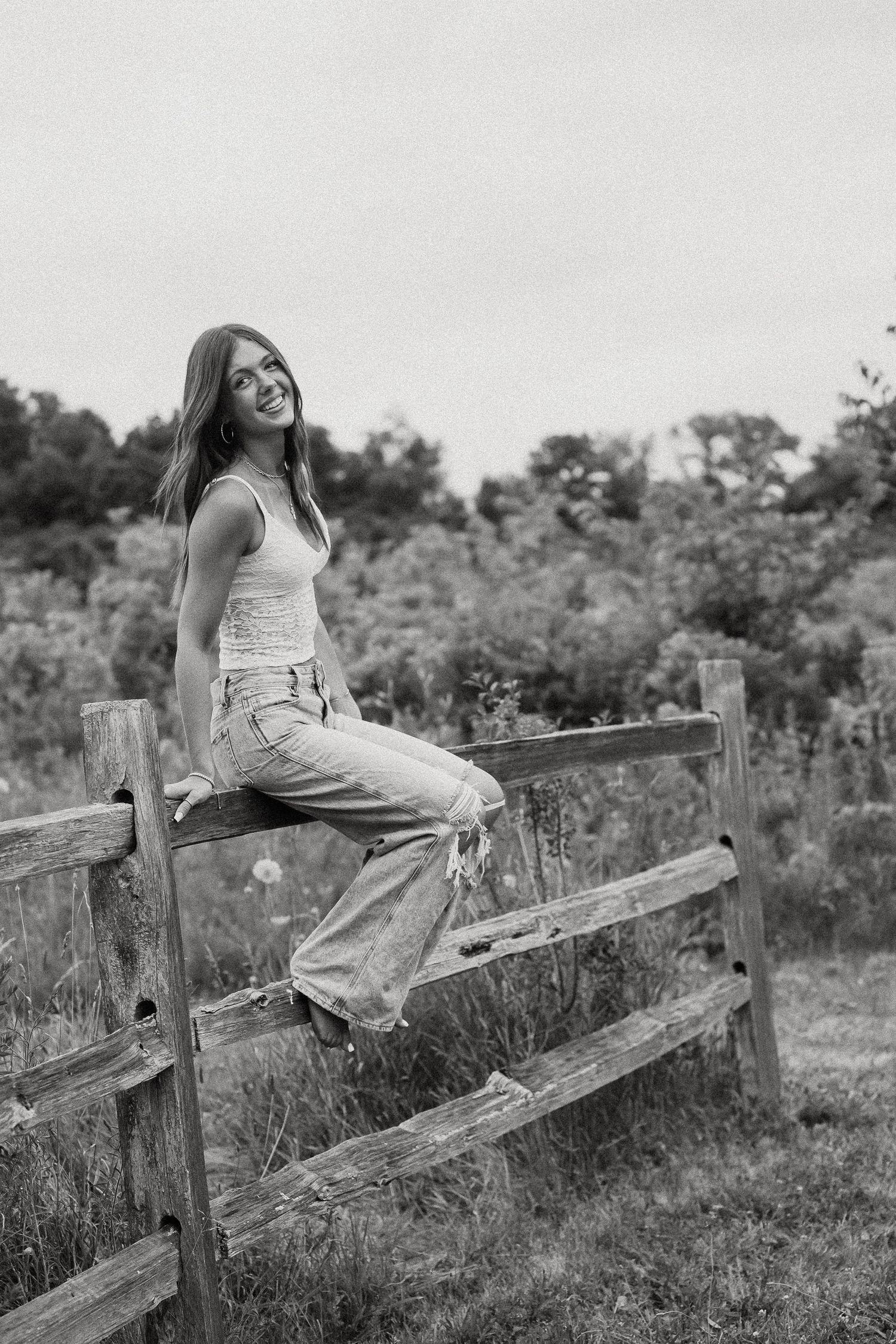 Smiling young woman in white tank and wide-leg jeans sitting on rustic wooden fence outdoors, black and white photo.
