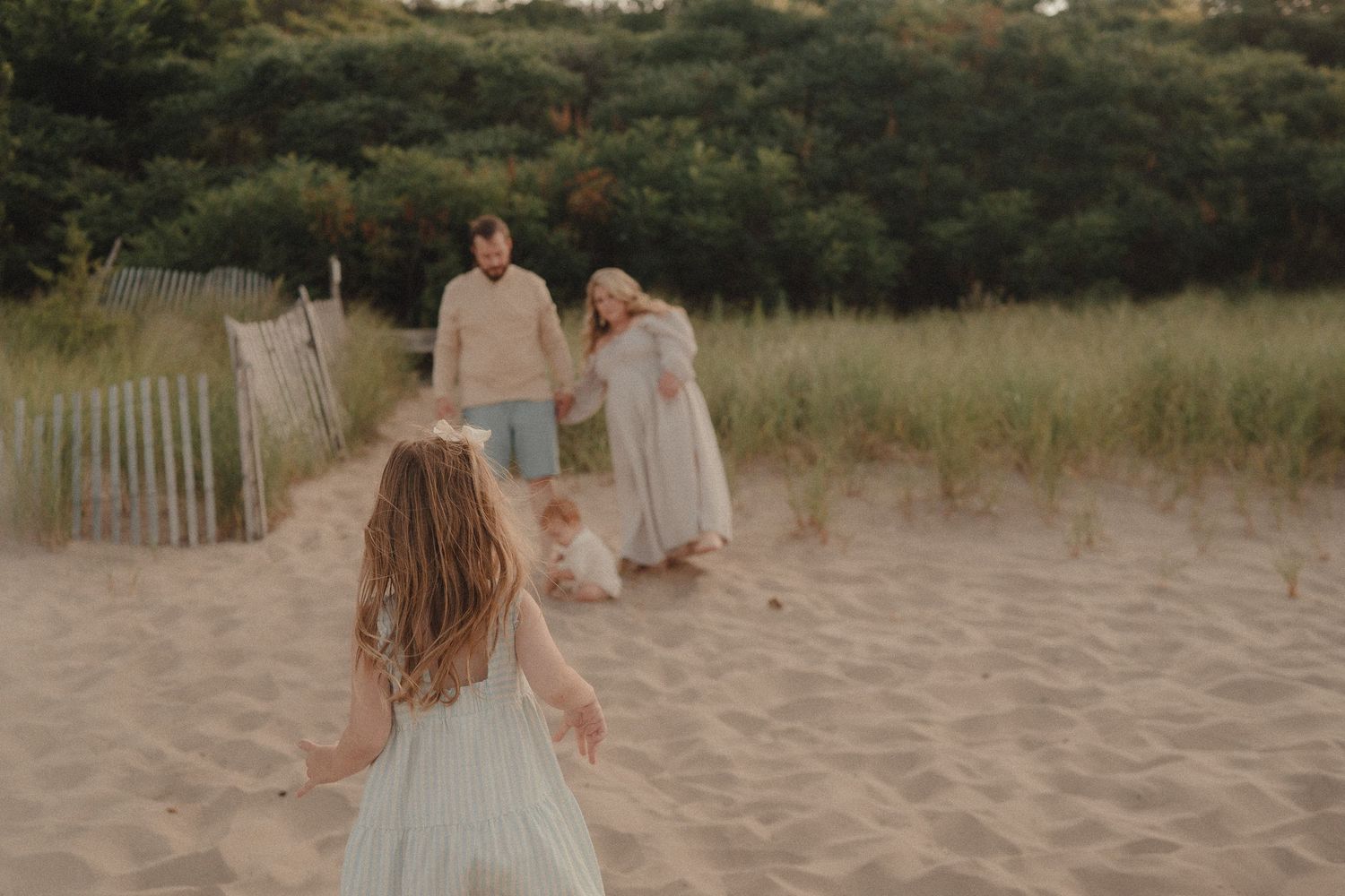 A young girl in a blue dress runs toward her parents and sibling on a sandy beach path lined with dunes.
