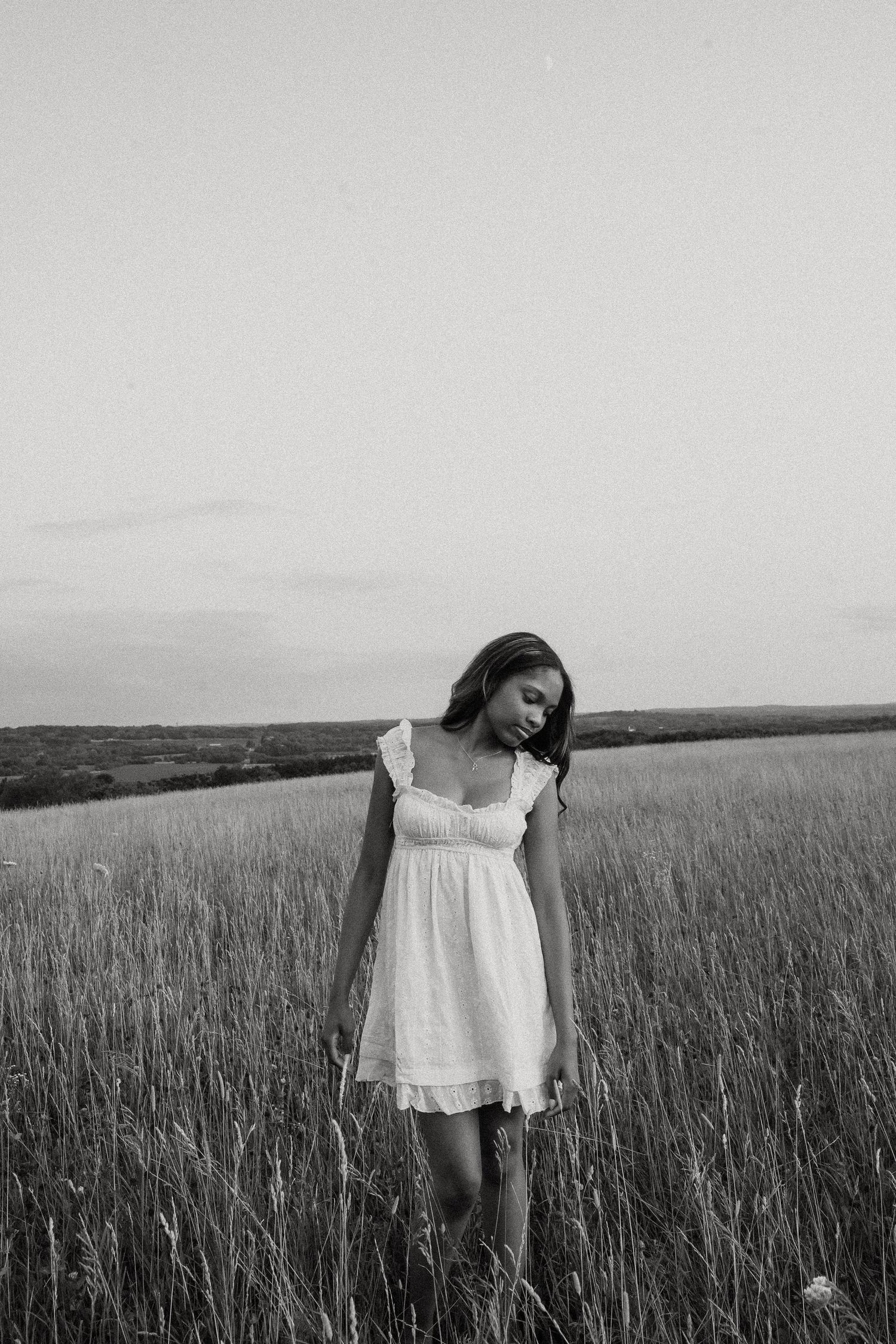 Black and white portrait of a young woman in a white dress standing in a vast open field.