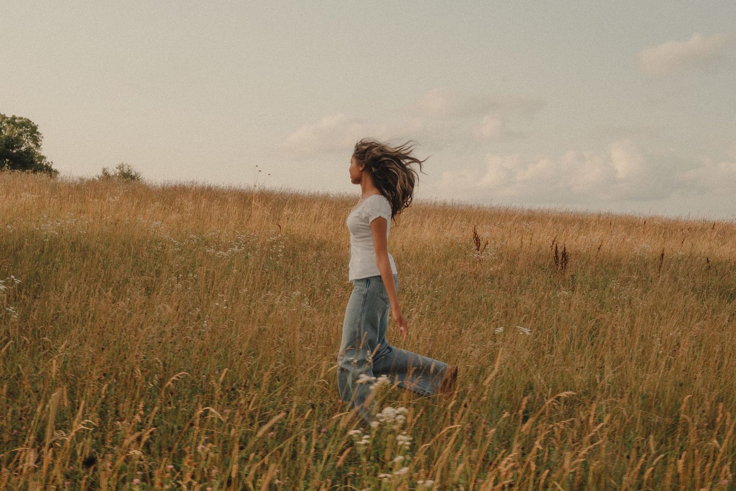 A young woman in jeans and white top runs freely through a golden meadow with wildflowers under a cloudy sky.