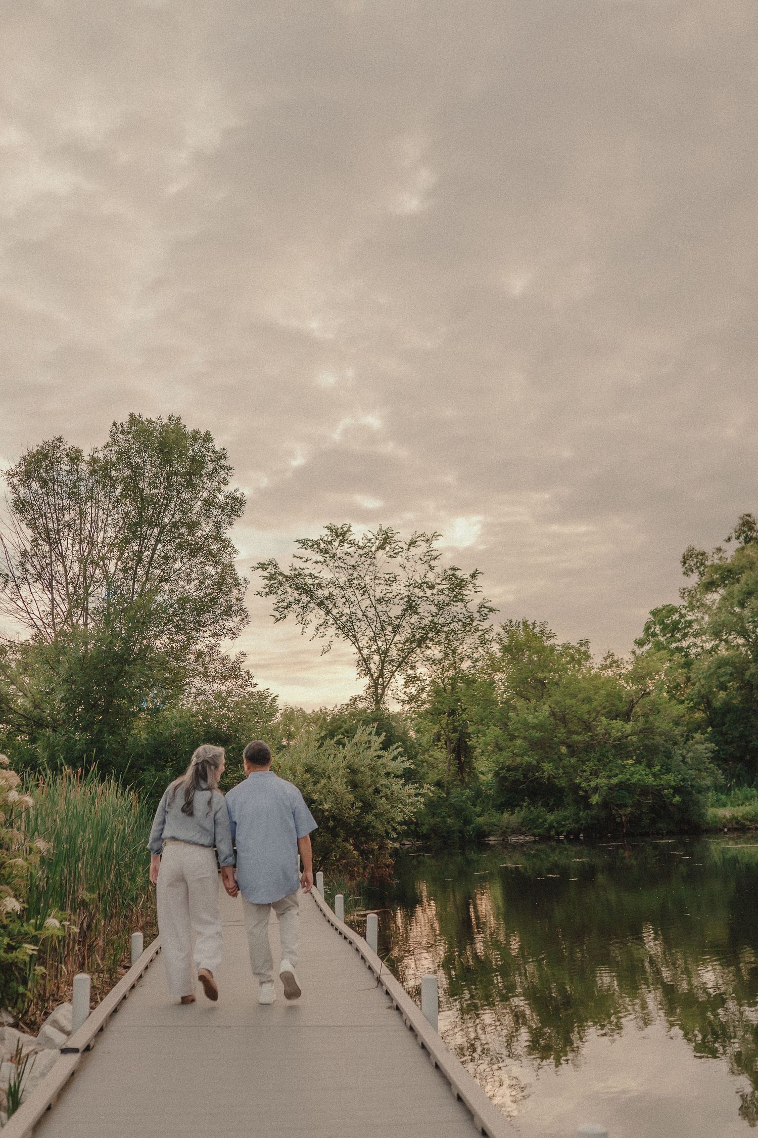 Couple holding hands walking along scenic wooden boardwalk beside calm reflective river at dusk.