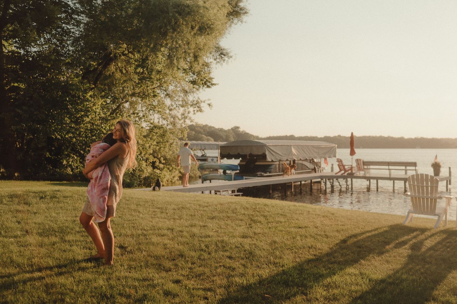 A young girl in a pink dress walks across a sunlit lawn near a lake dock.