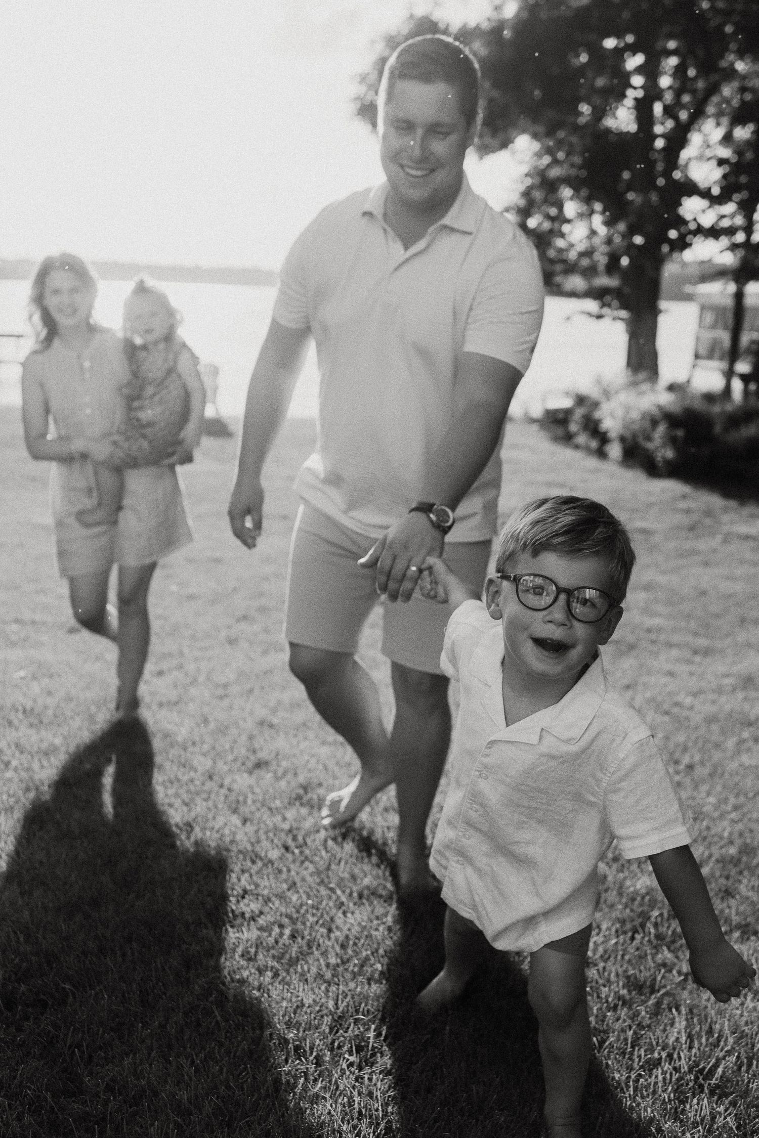 Black and white photo of a happy family walking on grass near a lake, with a smiling boy in glasses leading the way.