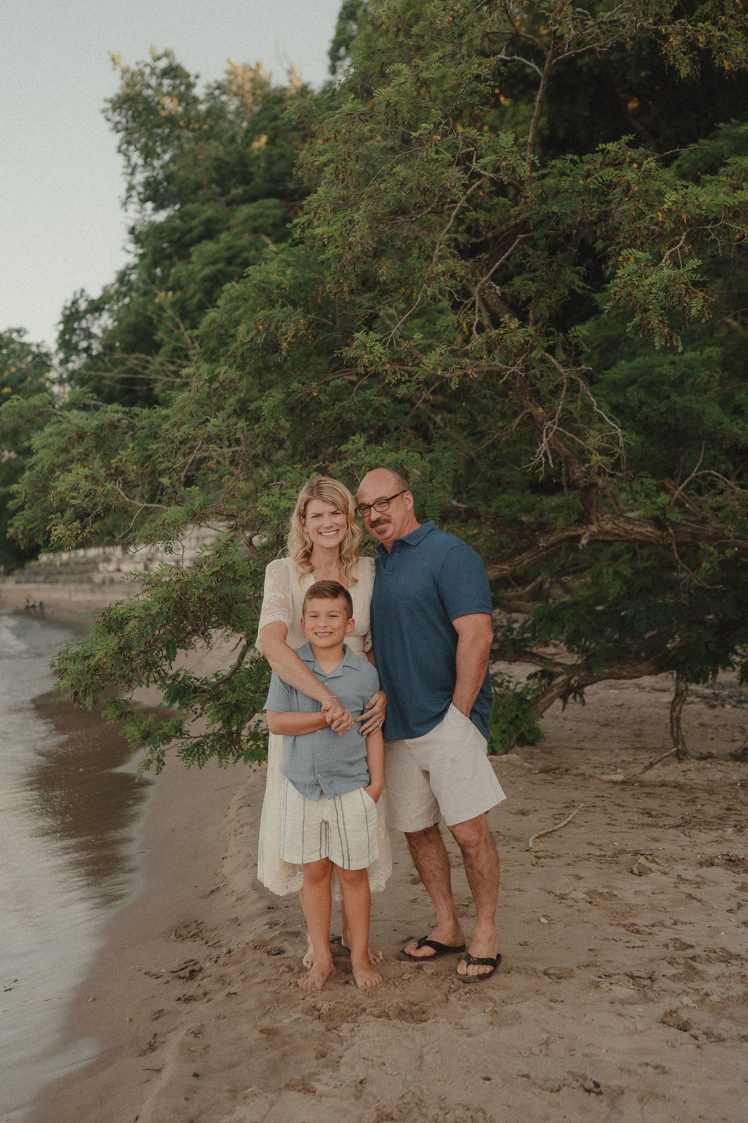 Family of three posing together on a sandy beach with lush green trees behind them.