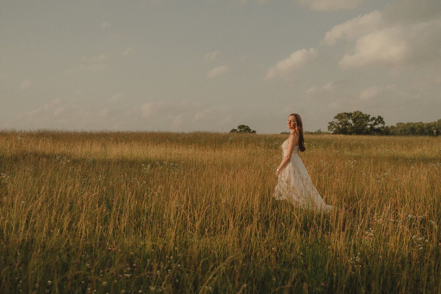 Young woman in white flowy dress standing alone in vast golden field under dramatic cloudy sky.
