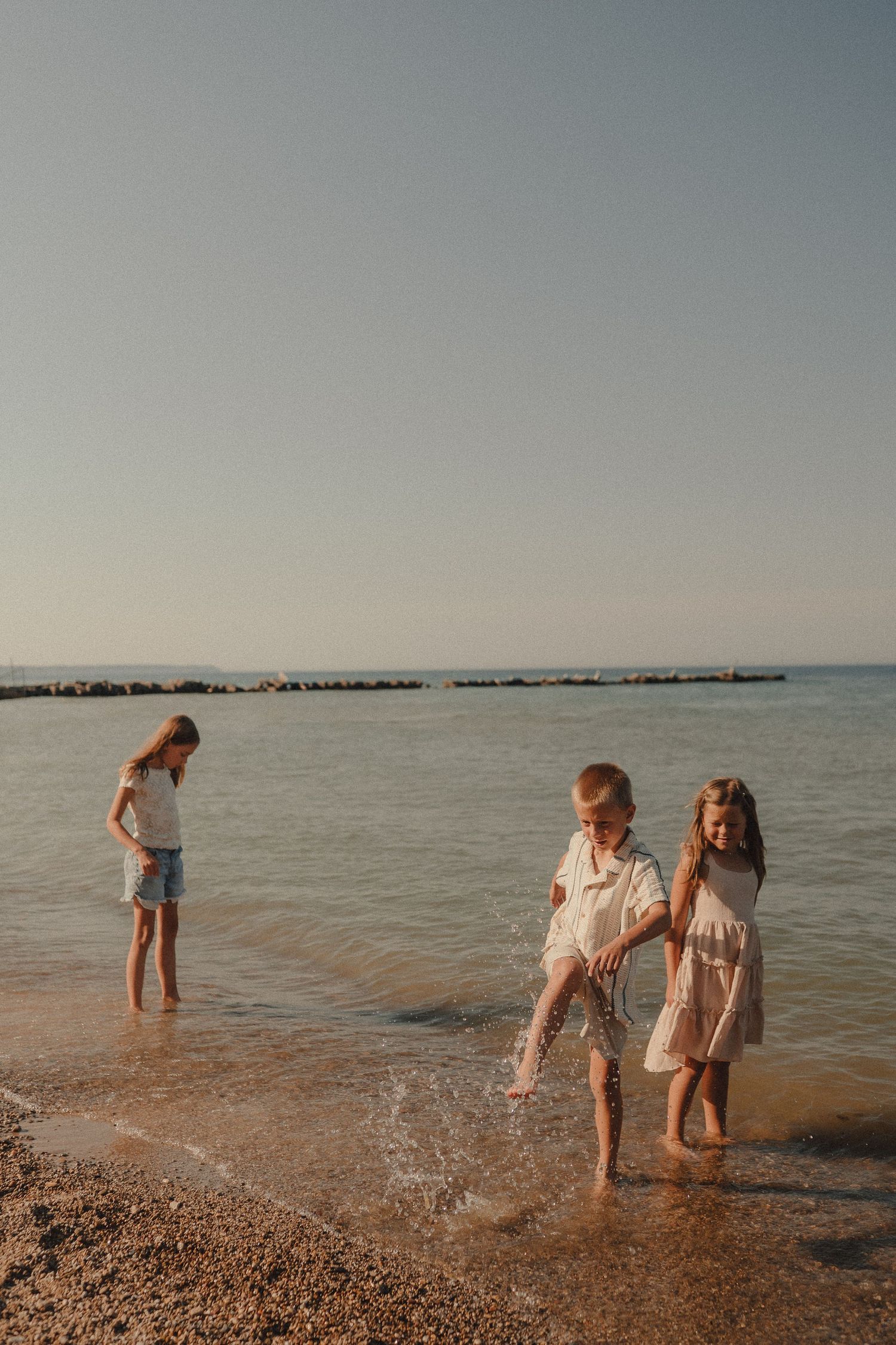 Three barefoot children splash and play in shallow ocean waves on a sunny beach during golden hour.
